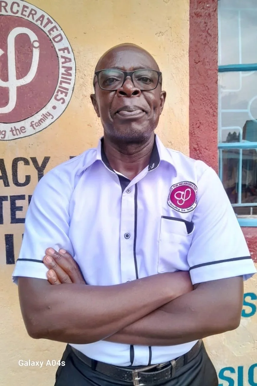 A man wearing glasses stands with his arms crossed looking directly at the camera while standing in front of a red and yellow painted building.