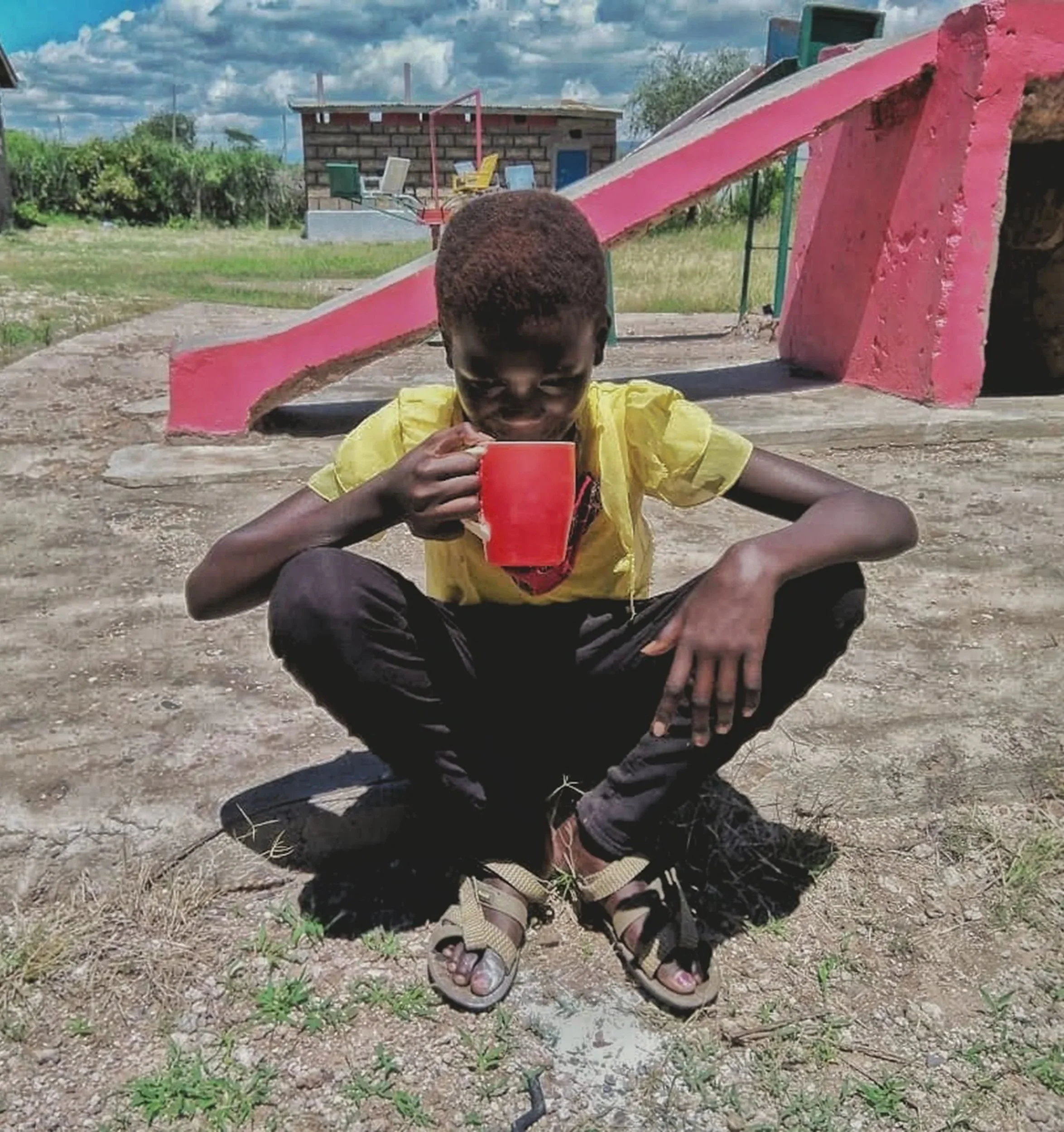 A child crouches on the ground outdoors, holding a red cup to their mouth, with a playground slide and a small building in the background.