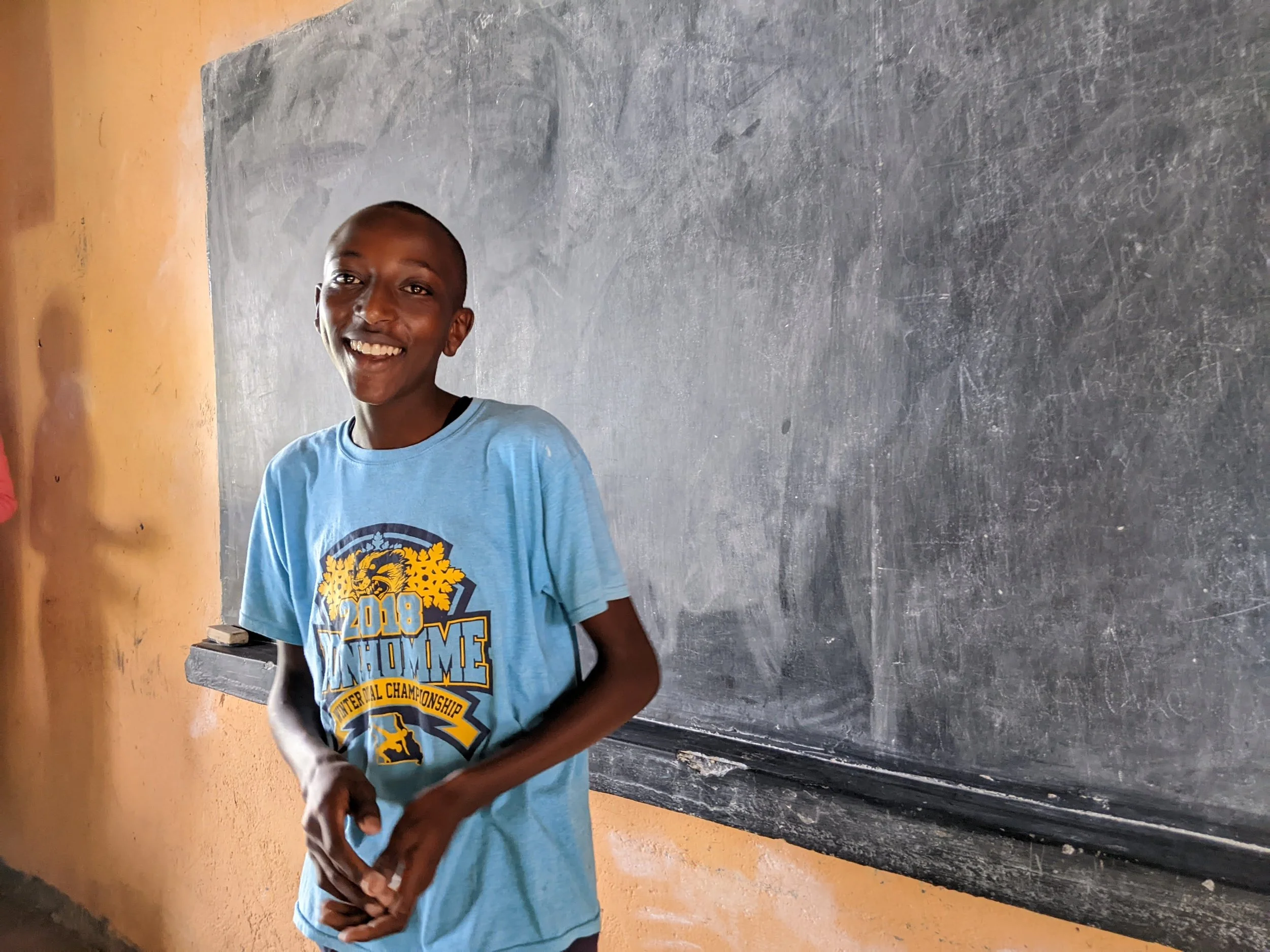 A teenage boy stands smiling in front of a chalkboard inside a classroom.
