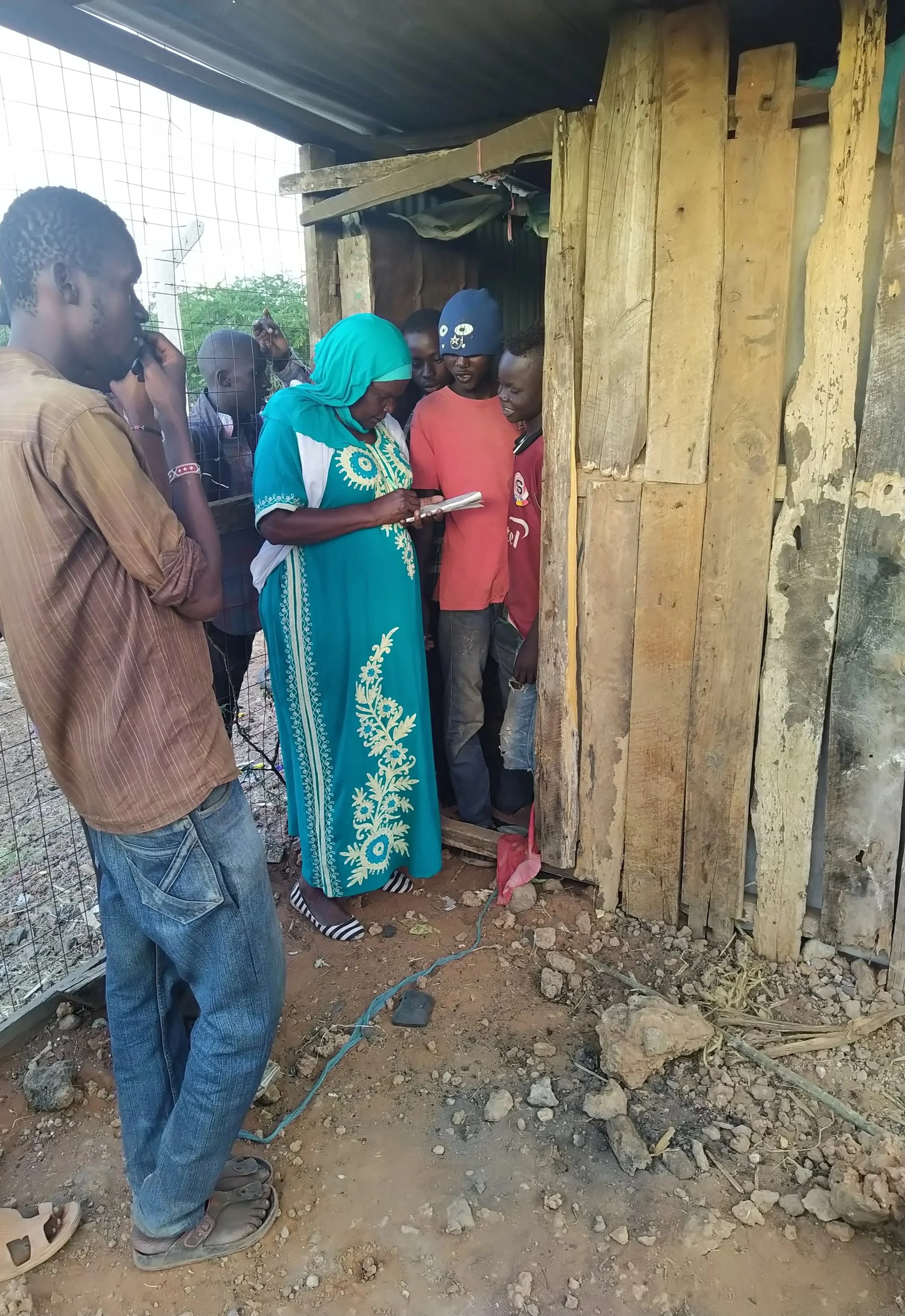 A group of male adults and teenagers stand closely together at the entrance of a small wooden structure, with an older woman wearing a headscarf writing in a notebook while others look on.