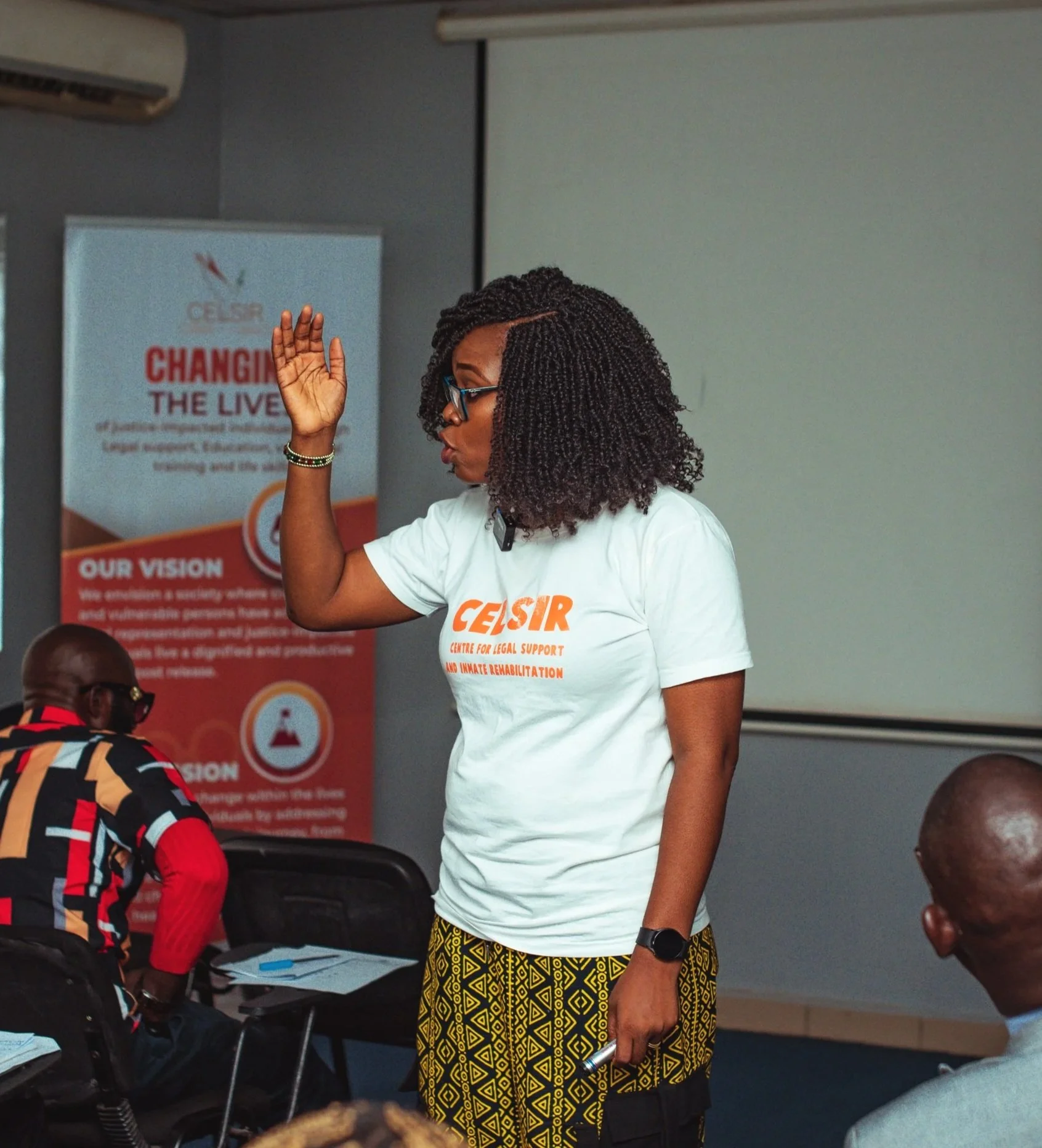 A woman wearing glasses gestures raises her hand while speaking in a workshop setting.