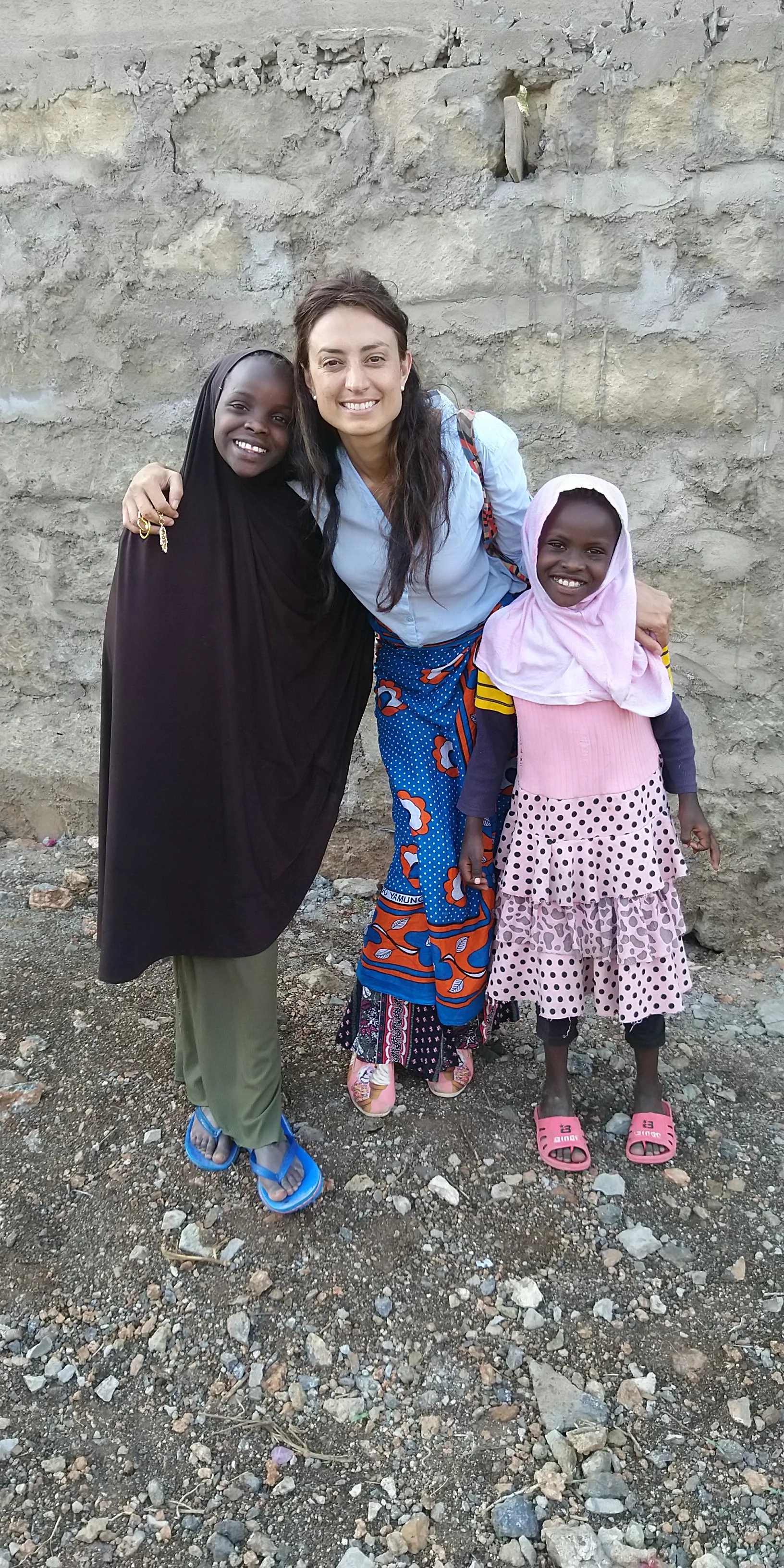 An adult woman and two children wearing headscarfs stand together smiling with their arms around each other outdoors, with a stone wall behind them.