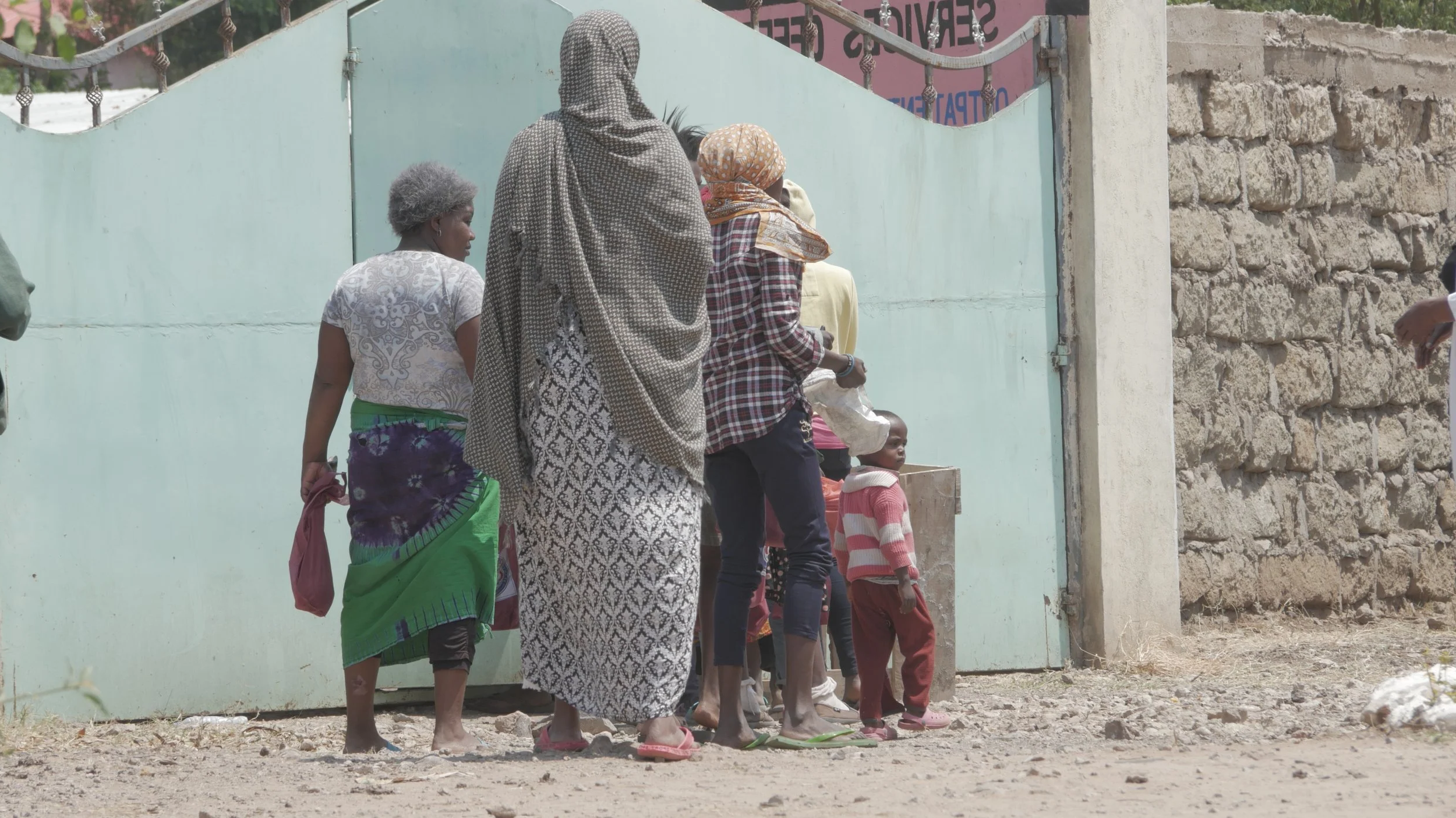 A small group of women and children stand near a large metal gate with door open, facing away from the camera.
