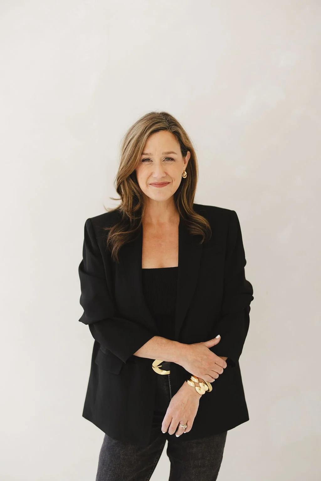 A woman with brown hair in a black blazer and top, standing against a plain white wall, smiling slightly with arms crossed.