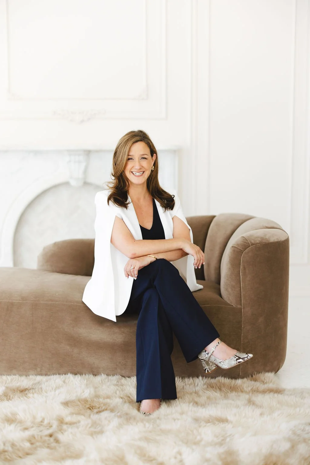 Smiling woman sitting on a beige sofa in a white room with a fireplace, wearing a black top, navy pants, white blazer, and snakeskin heels.
