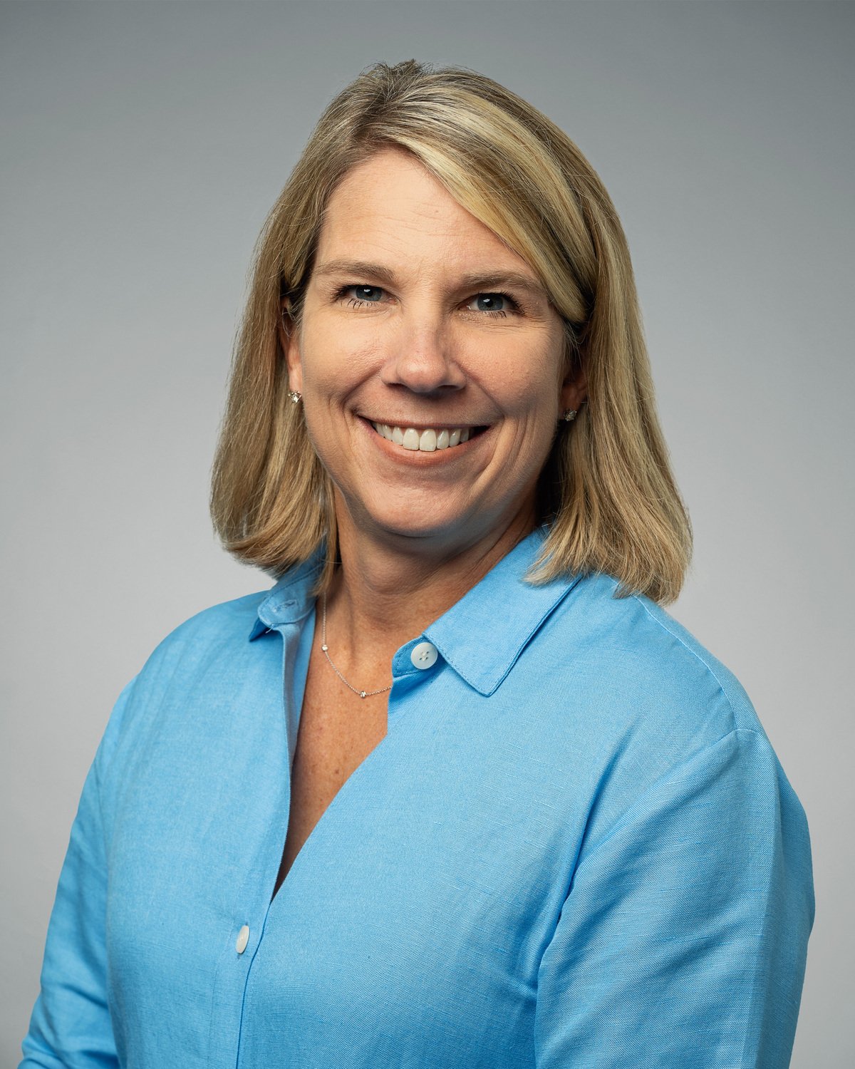 Portrait of a woman with blonde hair wearing a light blue shirt and small earrings, smiling against a plain gray background.