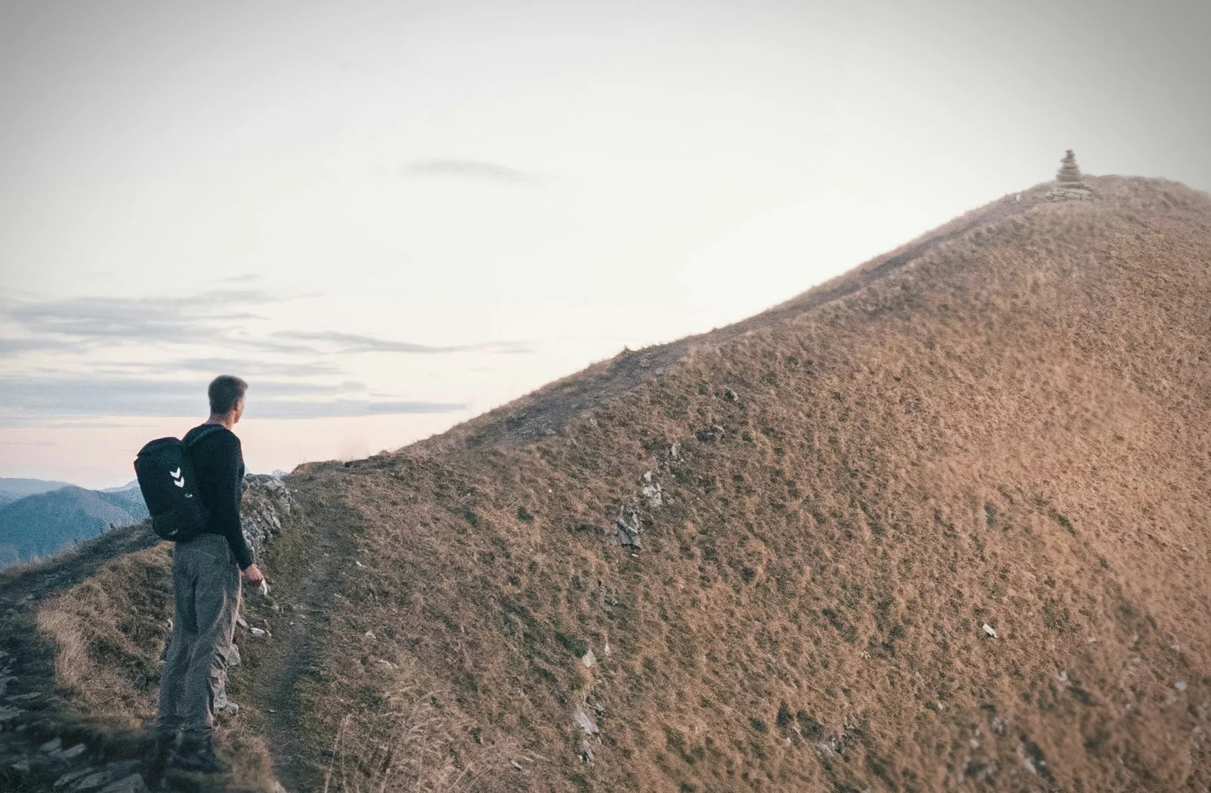 A person standing on a mountain trail, looking at a hill with a small pagoda on top, under a cloudy sky during sunset.
