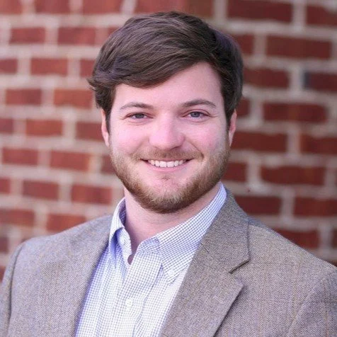 Portrait of a smiling young man with brown hair and a beard, wearing a gray blazer and a light blue shirt, standing in front of a brick wall.