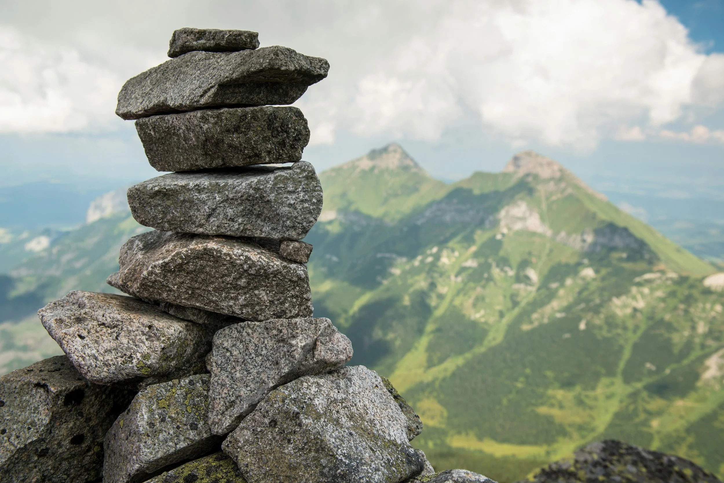Stacked rocks on a mountain overlooking green, rugged peaks and a blue sky with clouds.