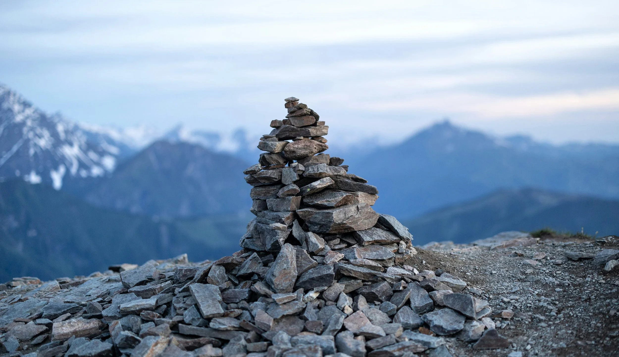 A stone cairn on a mountain ridge with snow-capped peaks and cloudy sky in the background.