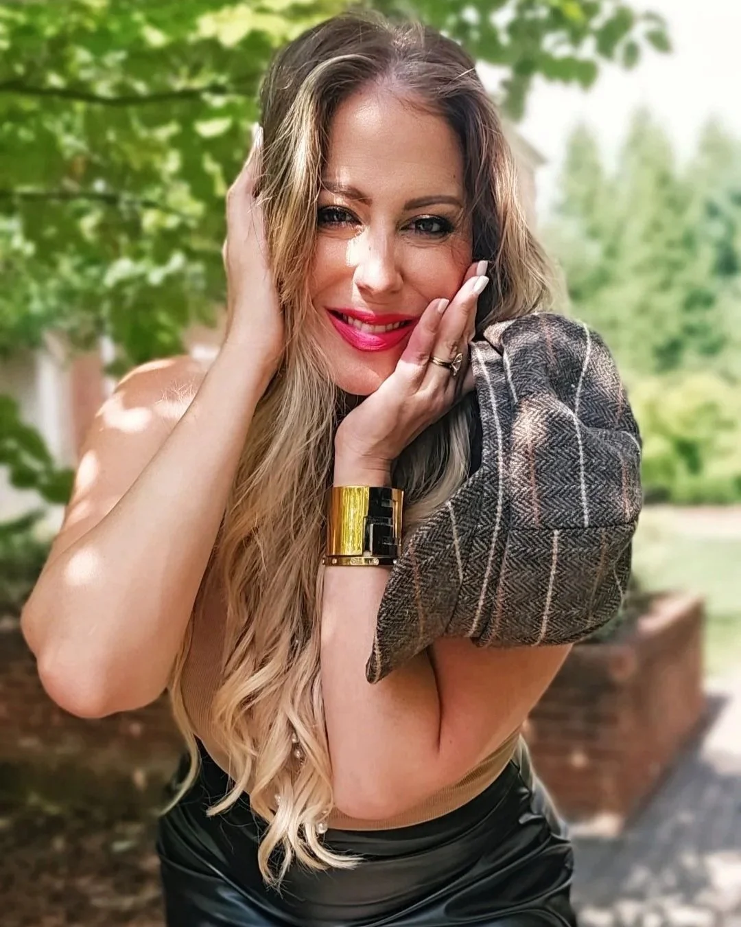 A woman with long blonde hair, pink lipstick, and a checkered blazer draped over her shoulder, smiling outdoors with a blurred green background.