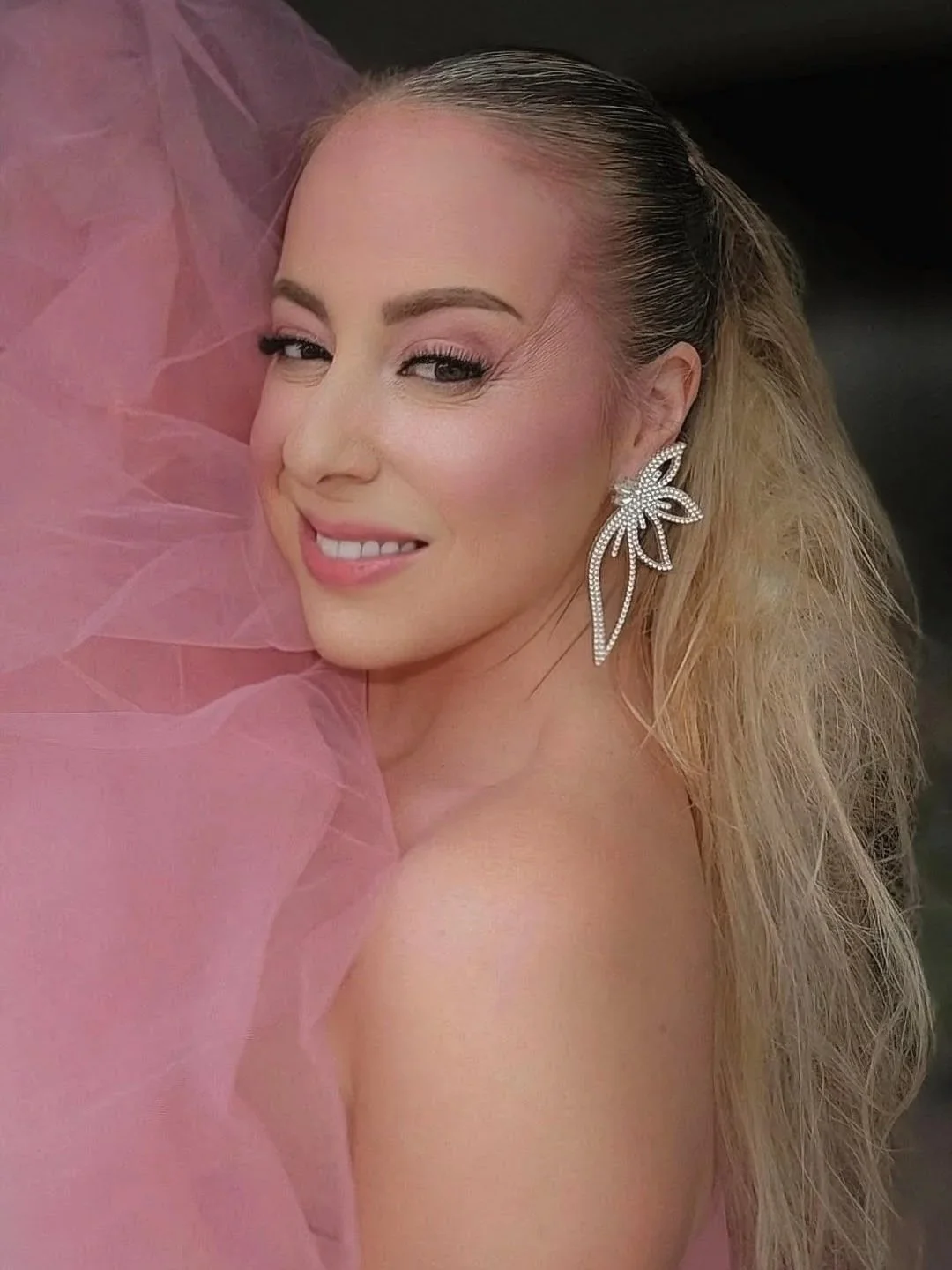 Close-up photo of a woman with blonde hair, light makeup, and large sparkling floral-shaped earrings, smiling softly and looking at the camera, with pink fabric in the foreground.
