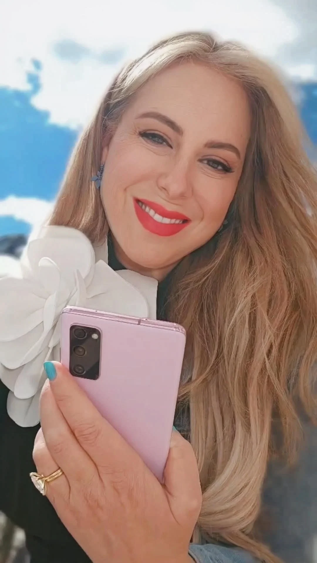 A woman with long blonde hair, red lipstick, and earrings is smiling while taking a selfie with a pink smartphone against a blue sky with white clouds.