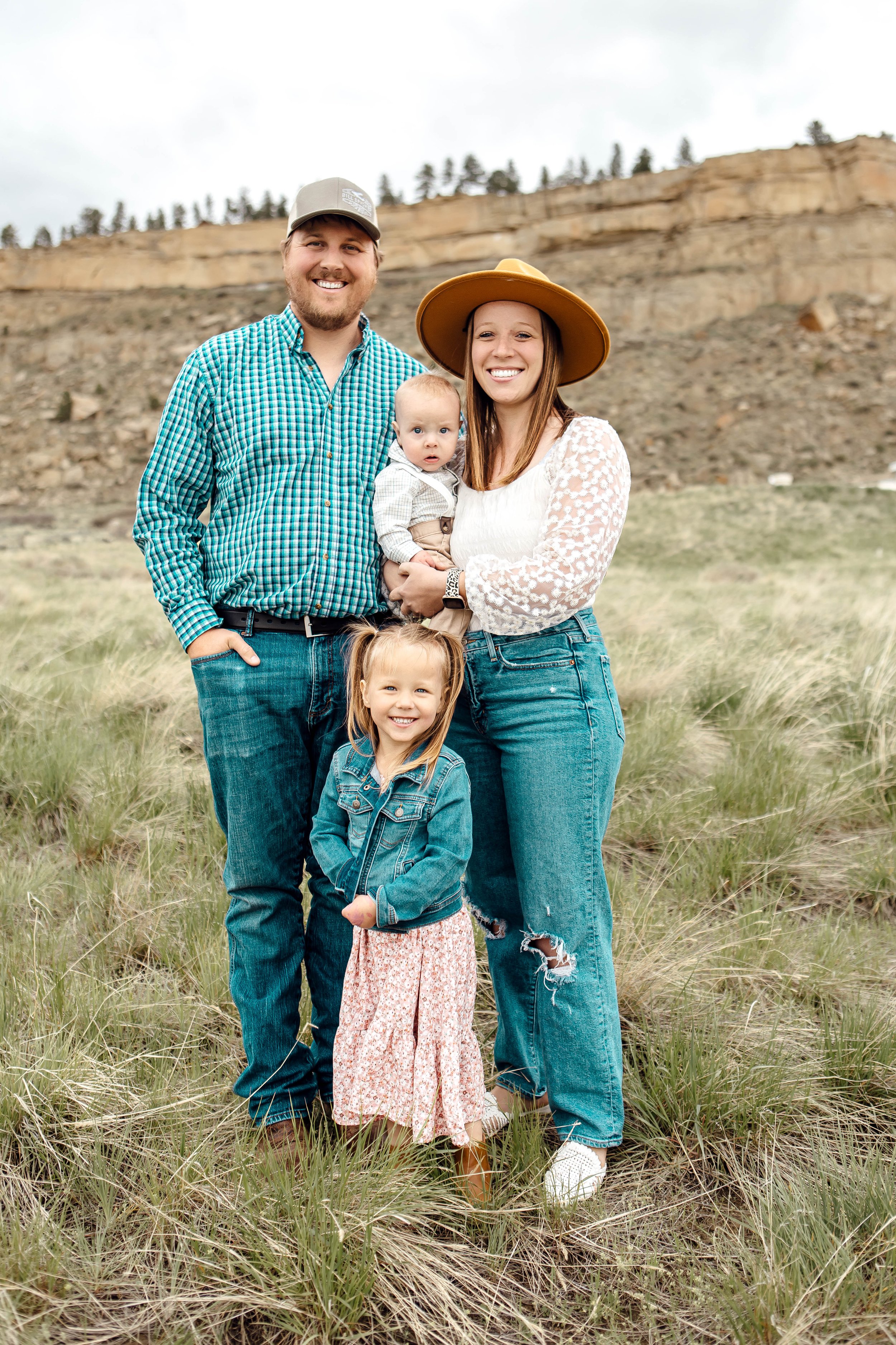 A happy family of four standing outdoors on grass with a rocky hill and trees in the background. The man is wearing a blue checkered shirt and a baseball cap. The woman is wearing a white lace top and a large yellow hat. The young girl is in a denim jacket and a patterned dress. The baby is being held by the woman and is wearing a beige sweater.