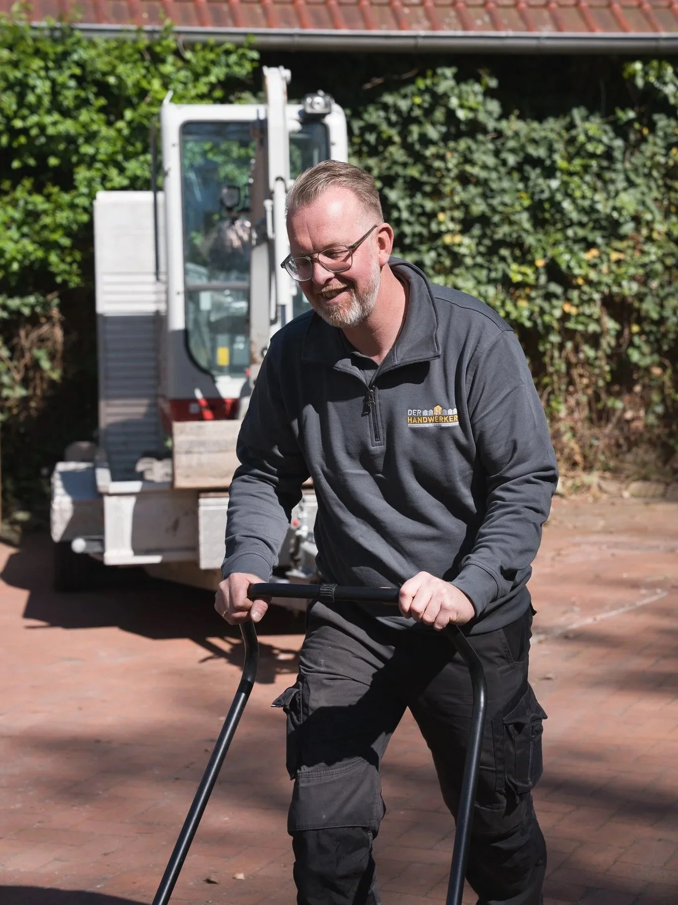 Ein Mann arbeitet im Freien an einem Stapel Holz, während er auf einem Fahrrad tritt, mit einem Klempner- oder Handwerker-Outfit und Brille, im Hintergrund ein Bauwagen.