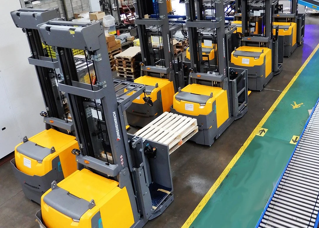 Row of yellow and gray Jungheinrich industrial forklifts parked in a warehouse, with a green and blue safety walkway on the right.