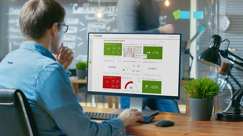 Man in glasses working at a desk with a computer monitor displaying manufacturing KPIs and data charts, newsroom with glass walls and plants in the background