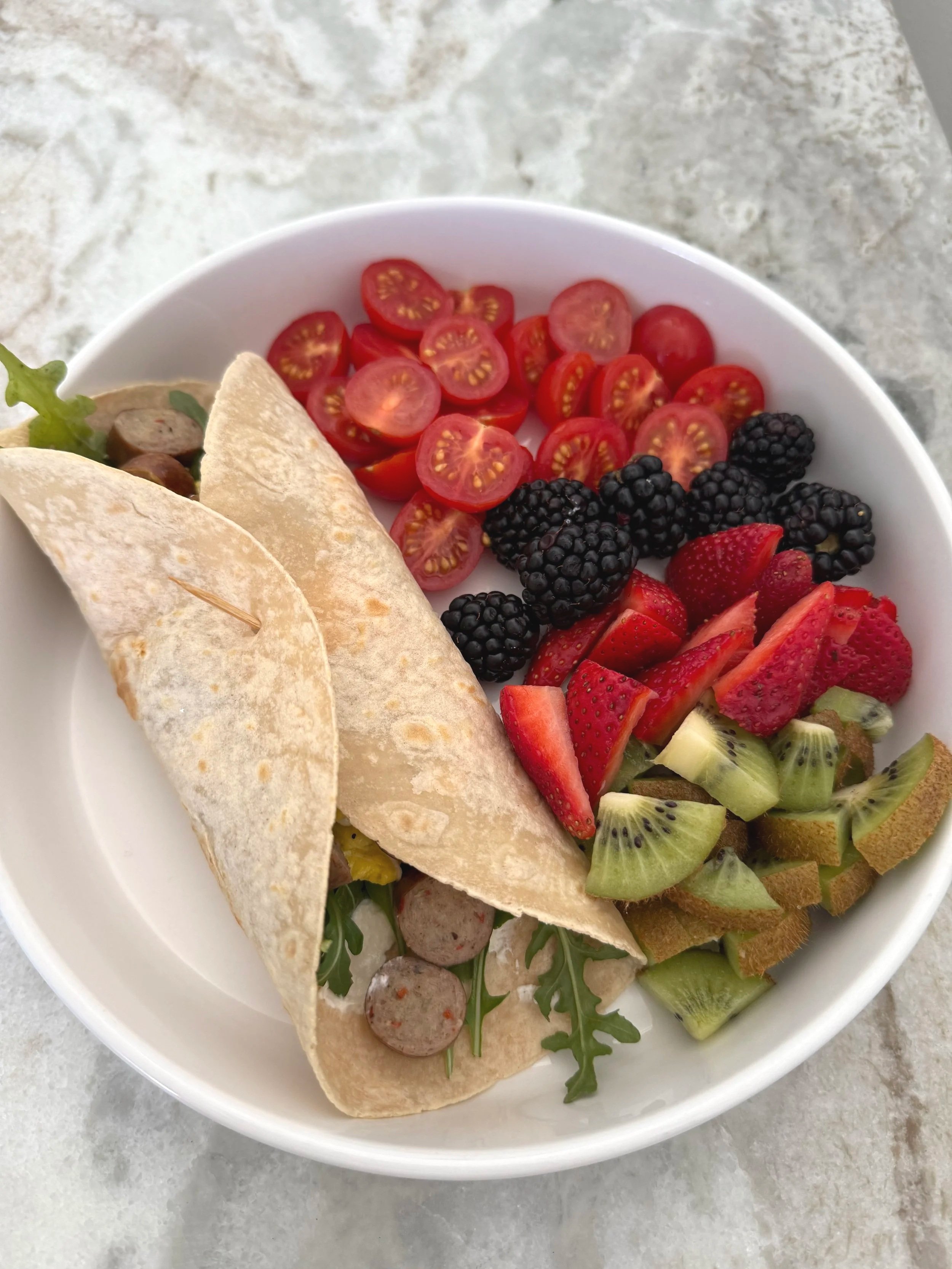 Bowl of fresh vegetables, strawberries, blackberries, cherry tomatoes, kiwi, and pita bread slices on a gray surface.
