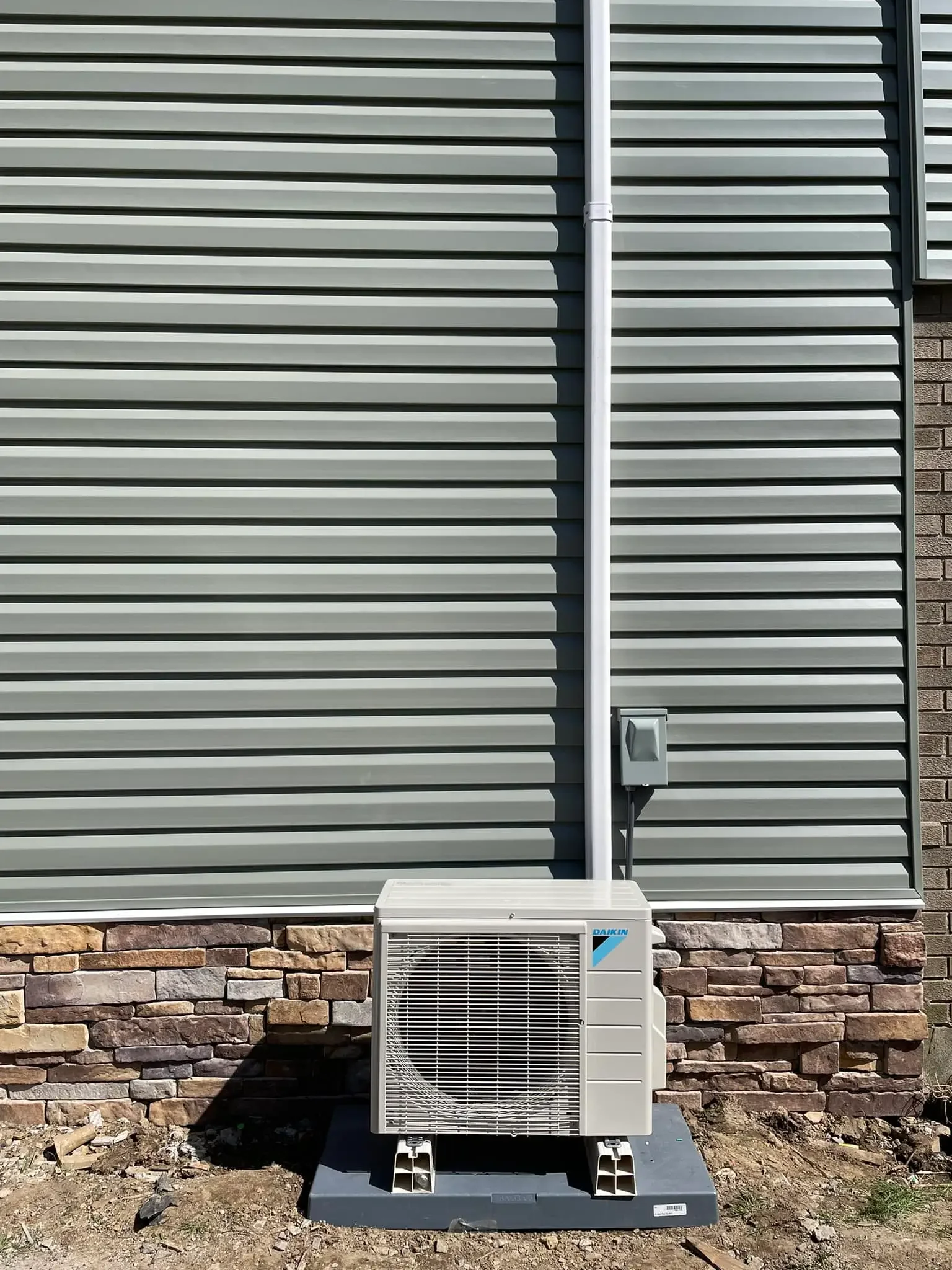 Exterior of a building with grey horizontal siding, a brick foundation, an electrical box mounted on the siding, and an outdoor air conditioning unit on a concrete pad.
