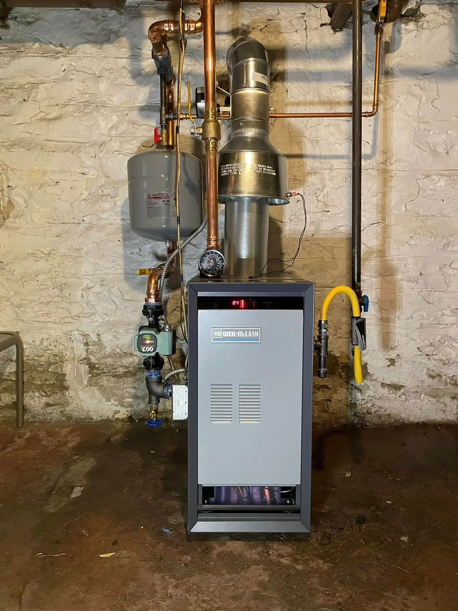 A boiler system with copper pipes, a gray water heater tank, a pressure gauge, and a control panel inside a basement with a stone wall and concrete floor.