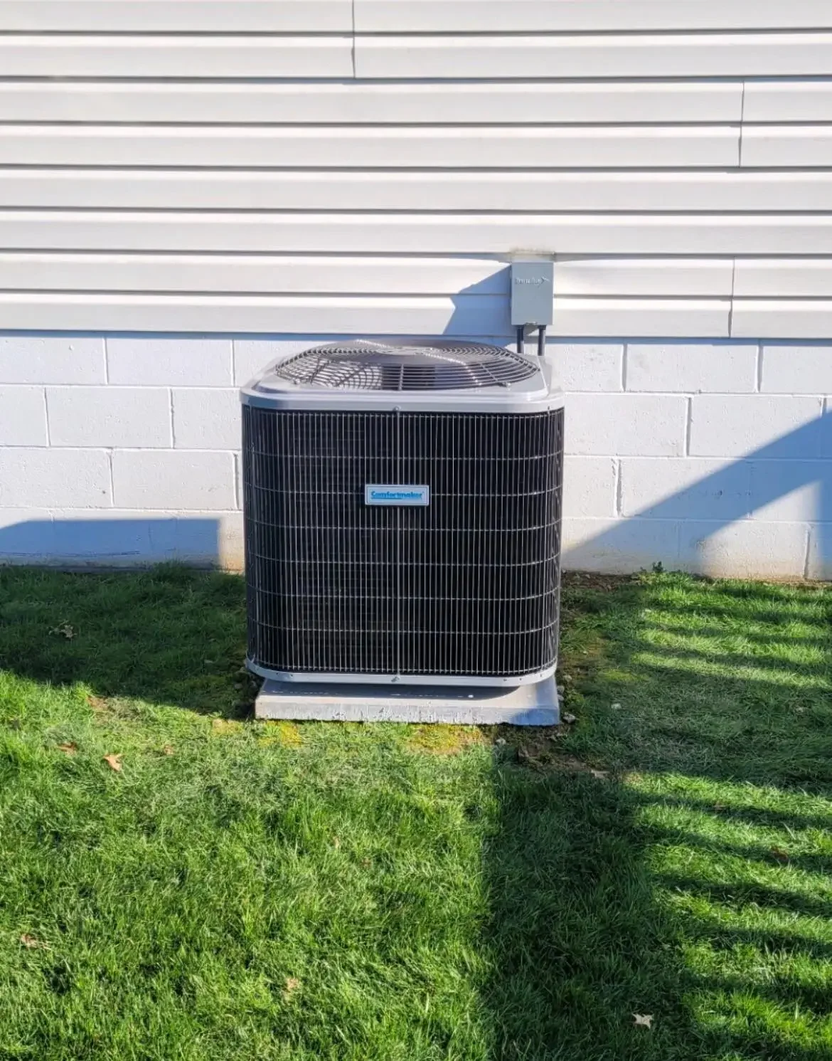 An outdoor air conditioning unit next to a white wall with vinyl siding, situated on a patch of green grass.