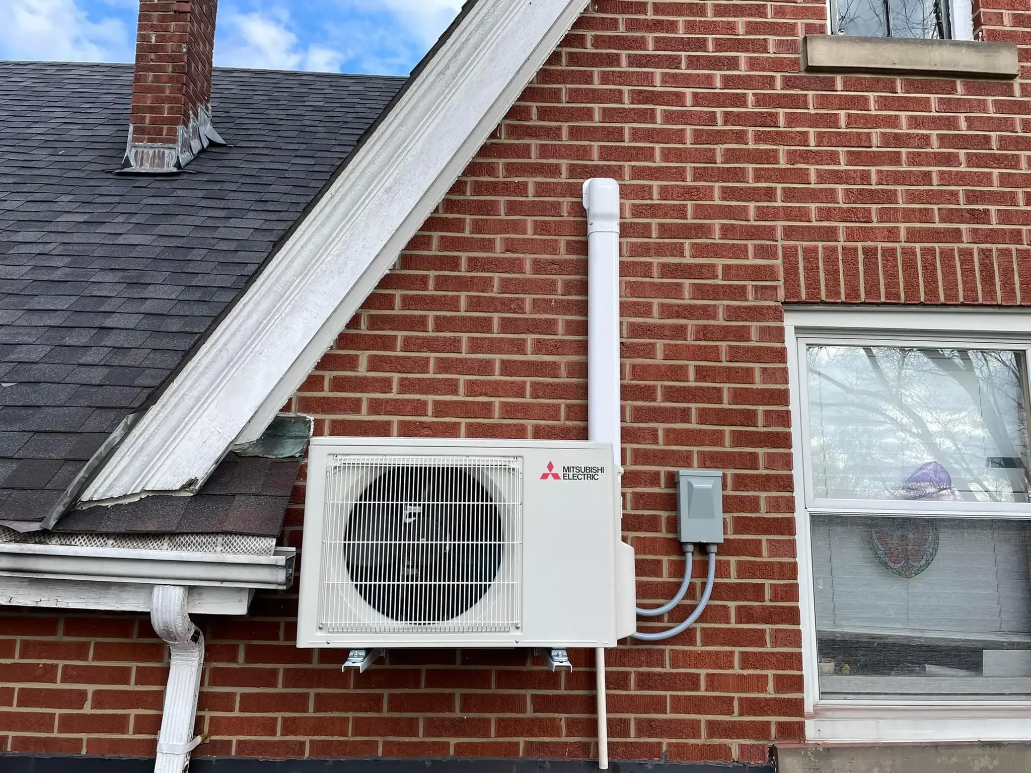 Exterior of a brick house with a Mitsubishi Electric heat pump unit mounted on the wall near a window and an outdoor vent pipe.