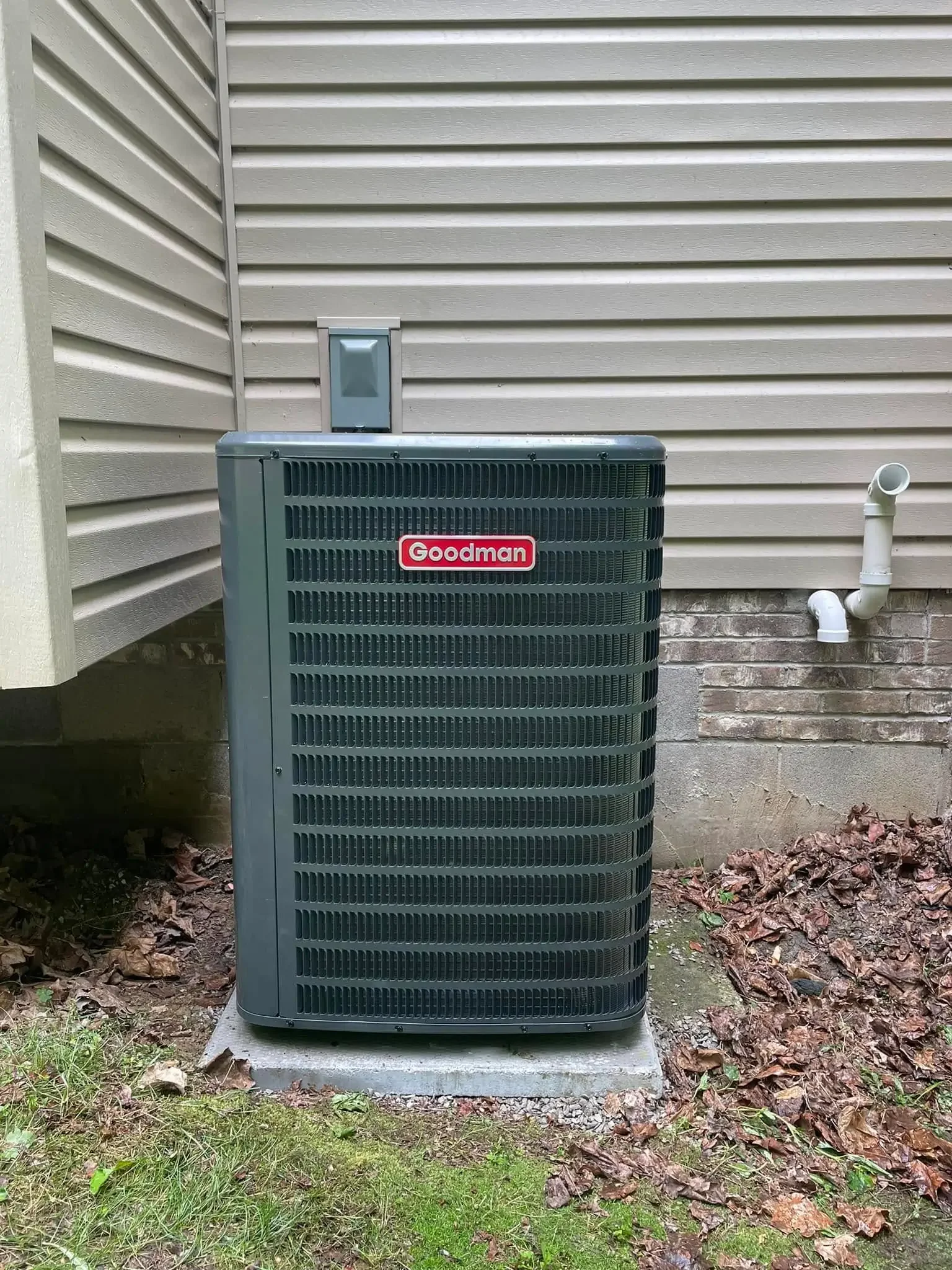 Exterior of a Goodman air conditioning unit installed on a concrete slab outside a house, with siding walls, a vent pipe, and some fallen leaves on the ground.