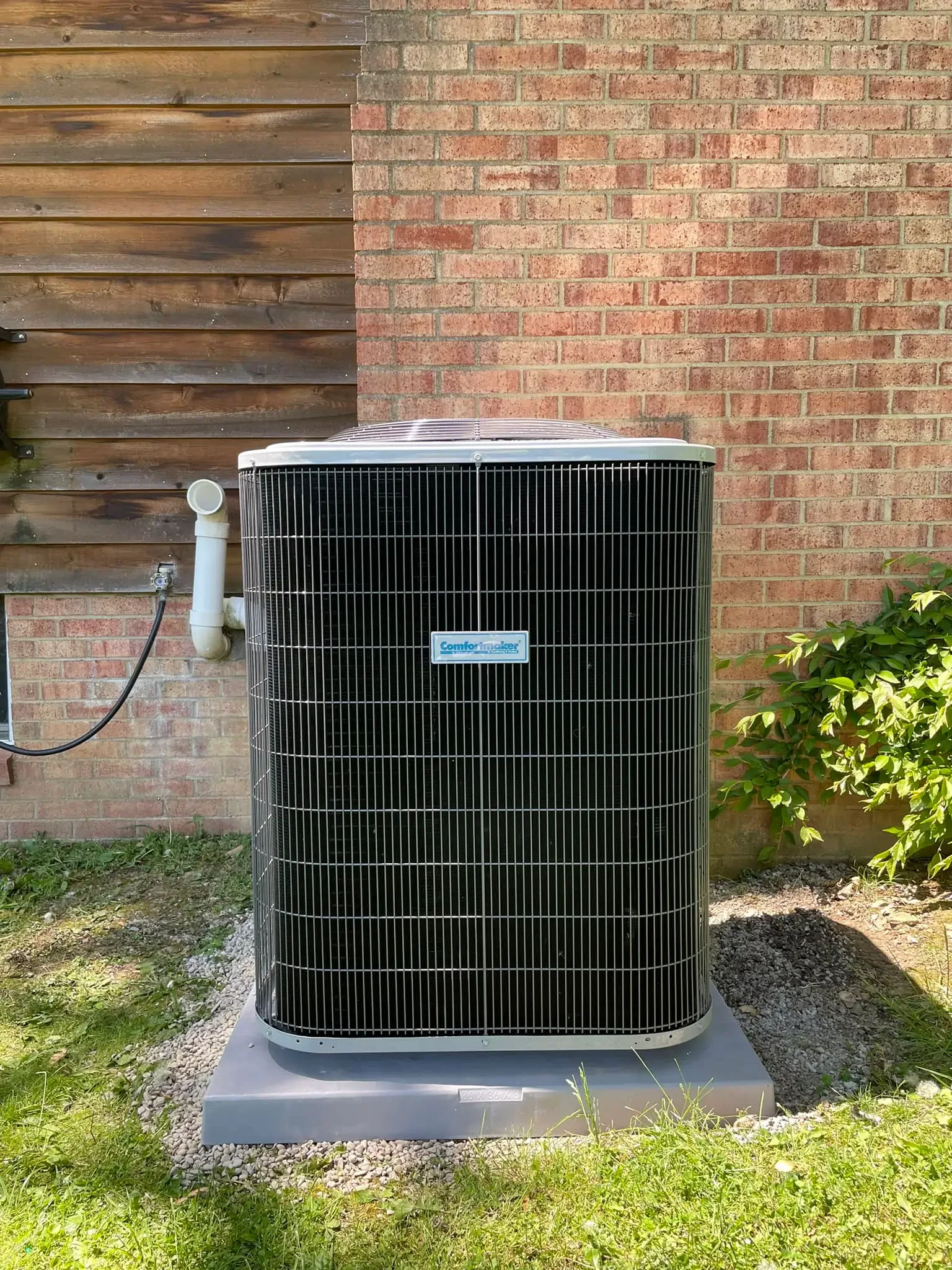 Outdoor air conditioning unit installed on a concrete slab, next to a brick wall and a wooden fence, with some greenery on the side.