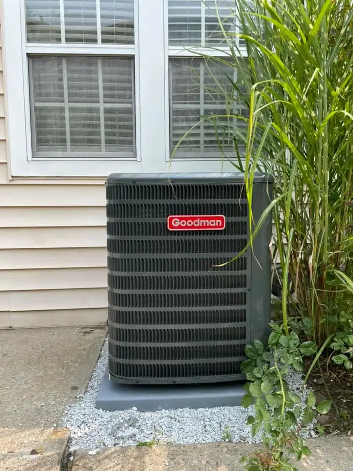 Central air conditioning unit outside a house near a window, with green plants and shrubs nearby.