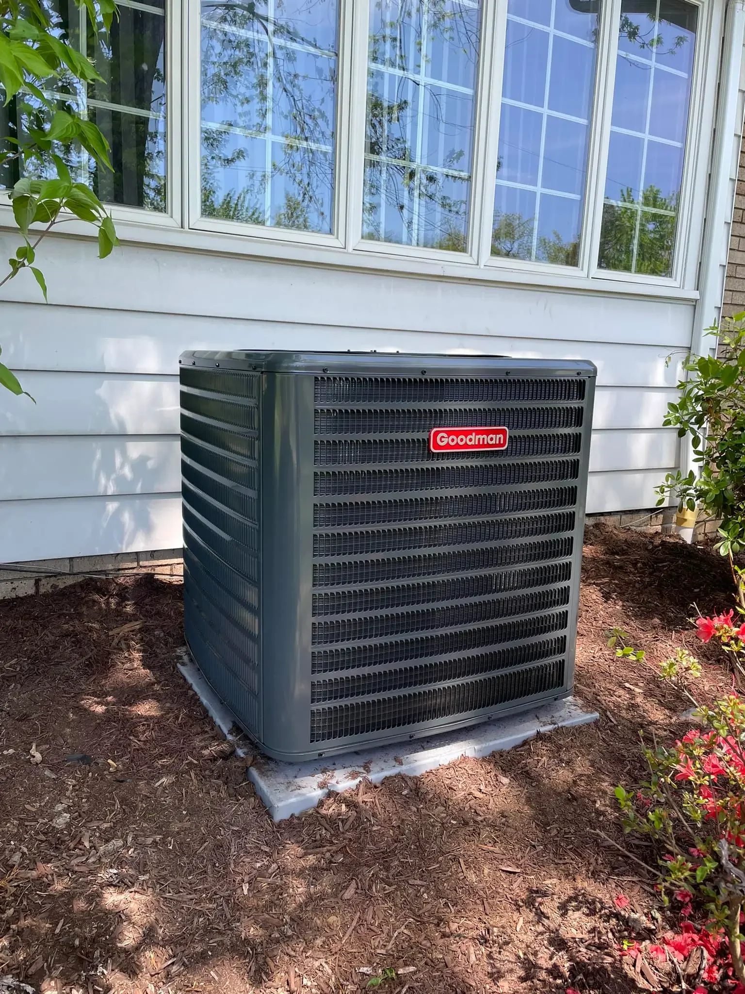 An outdoor air conditioning unit with a black grille and a red Goodman logo, installed on a concrete pad beside the white siding house and a window.