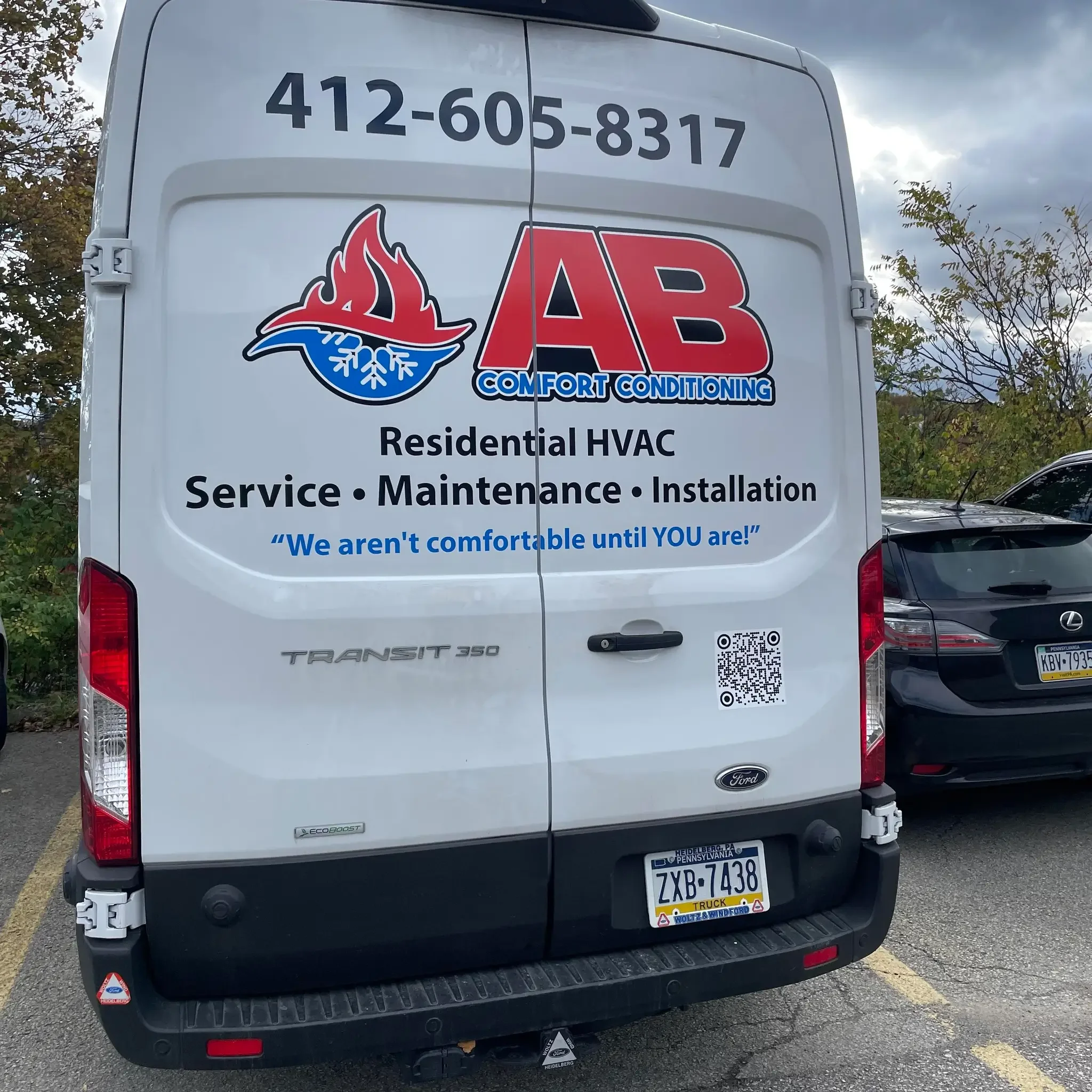 The back of a white Ford Transit 350 van with business information for AB Comfort Conditioning, an HVAC service company. The van displays the company's phone number, logo, services offered like residential HVAC, service, maintenance, and installation, along with the slogan "We aren't comfortable until YOU are!" There are also QR code and license plate details visible.