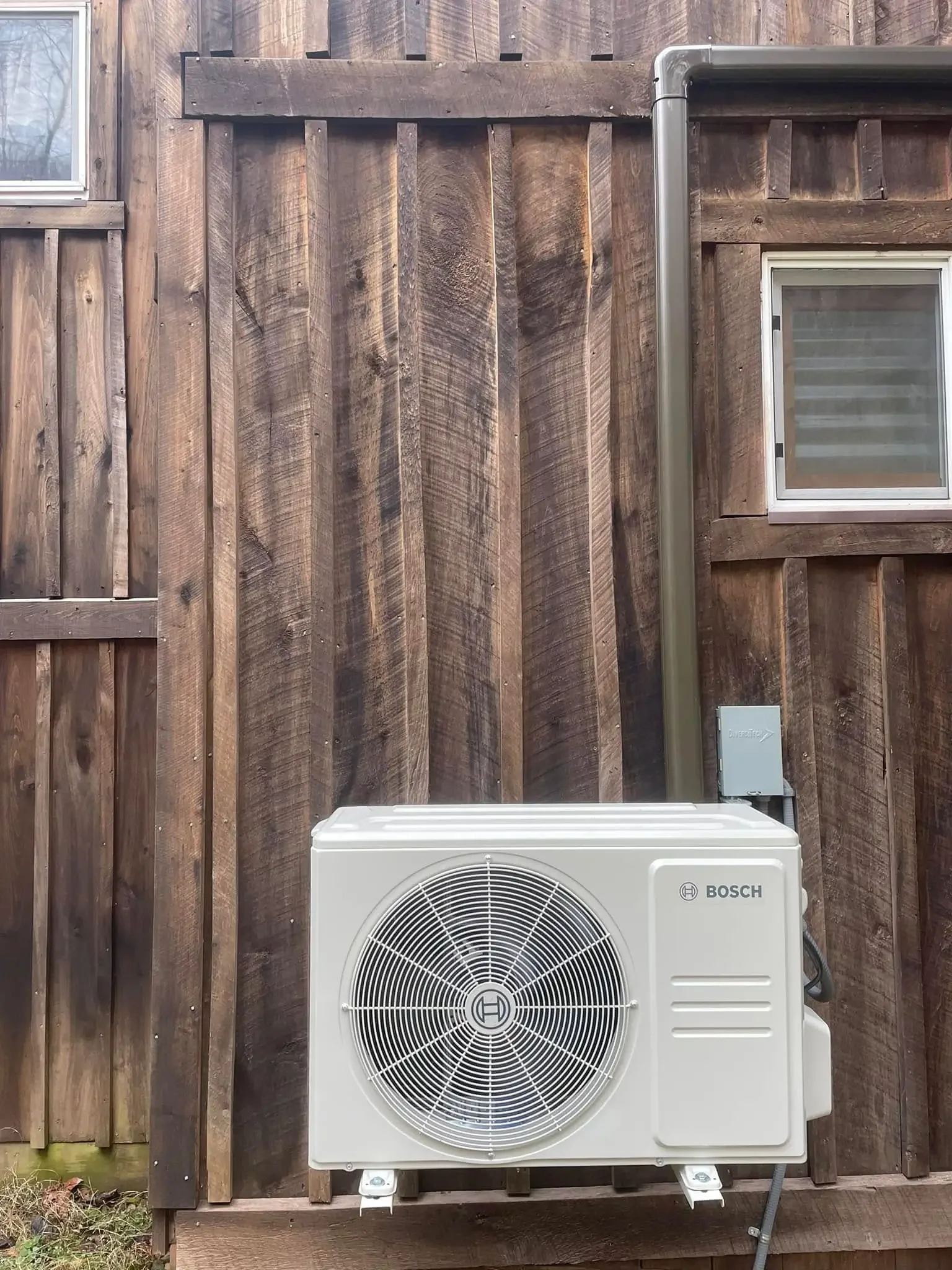 Outdoor air conditioning unit mounted on a wooden wall with a window and rain gutter.