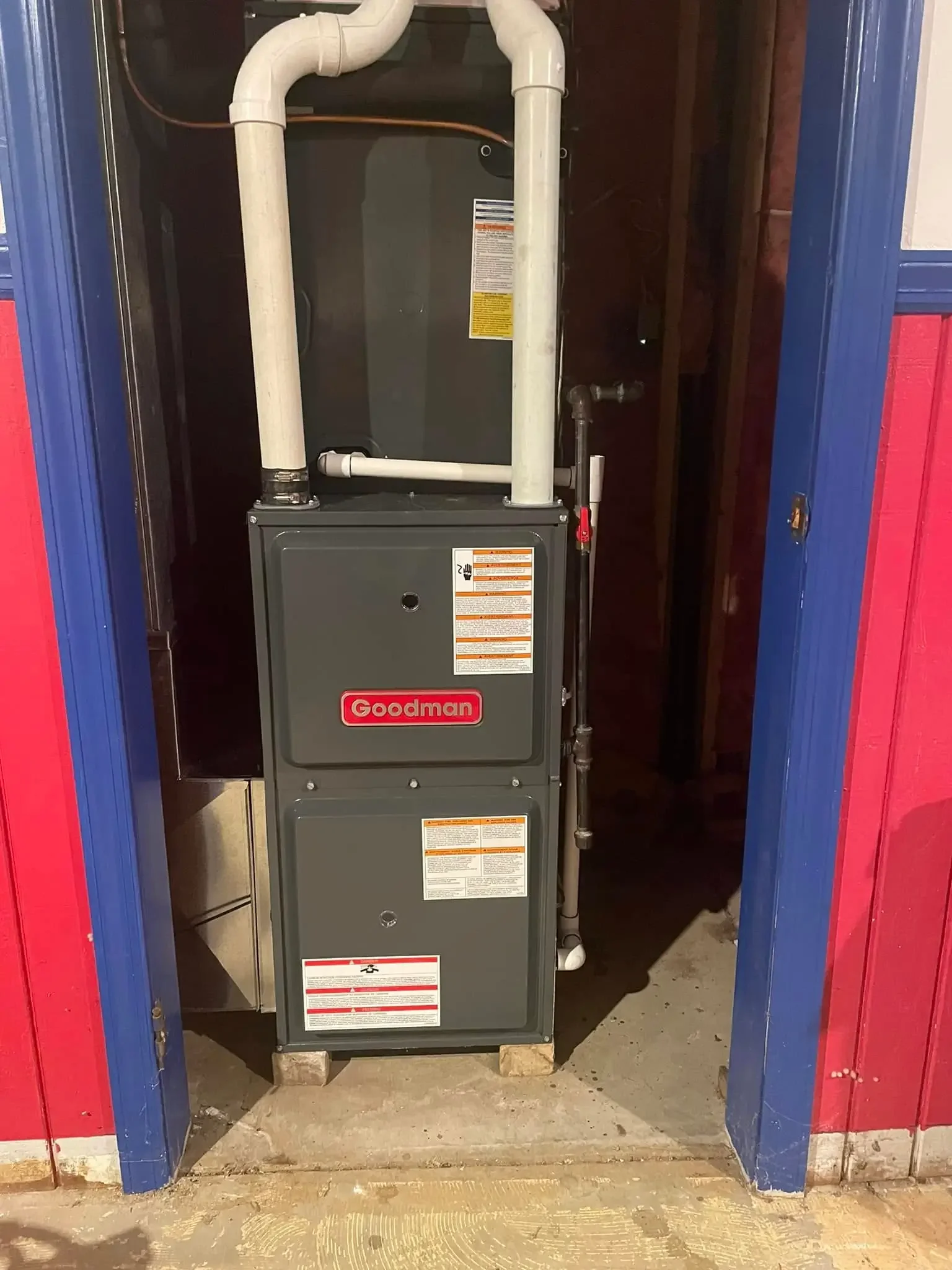 A Goodman furnace installation in a garage or utility space, with white PVC pipes connected to the top and various warning and instruction labels on the front.