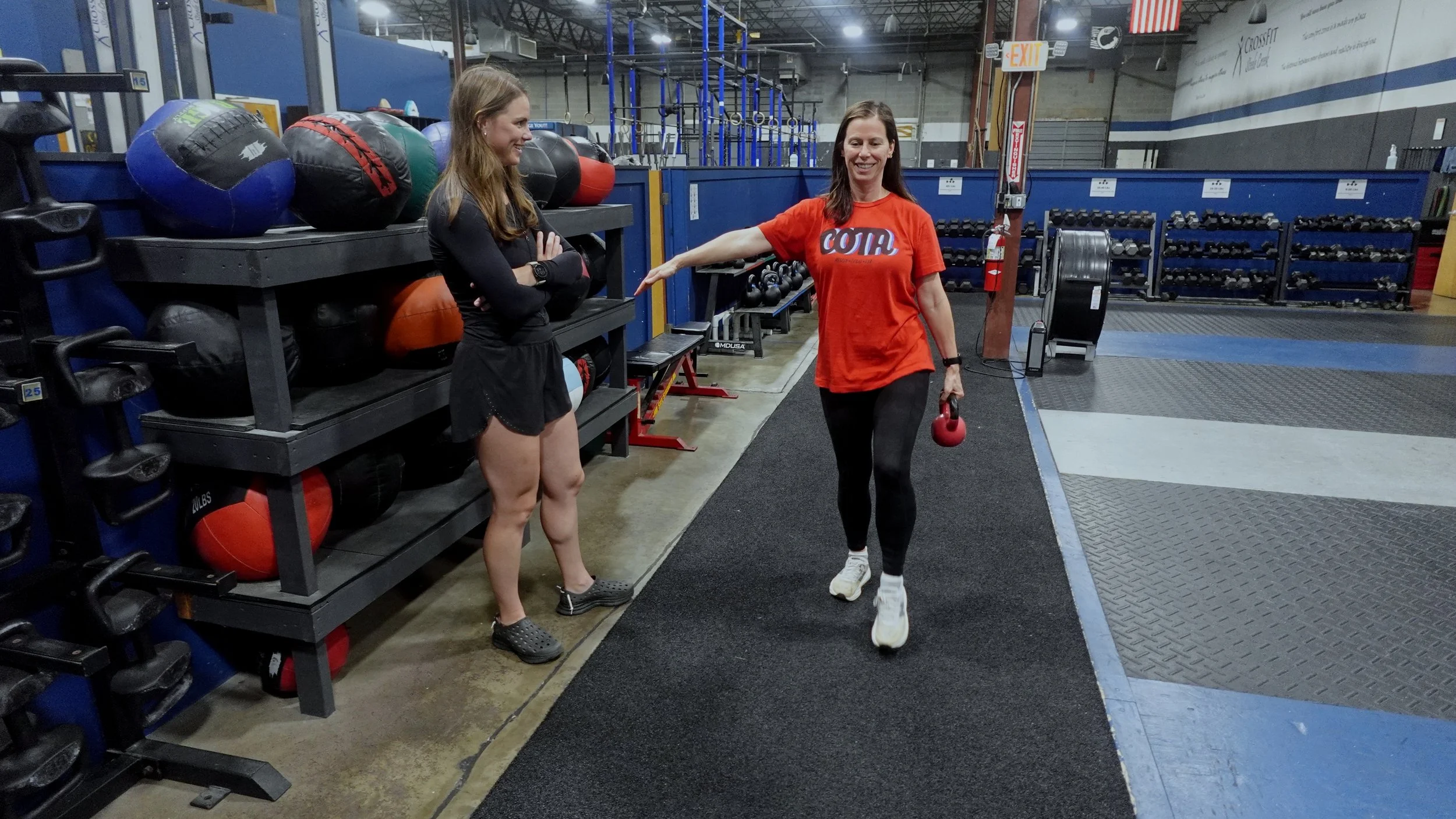 Two women at a gym, one holding a kettlebell and the other smiling, with gym equipment and weights in the background.