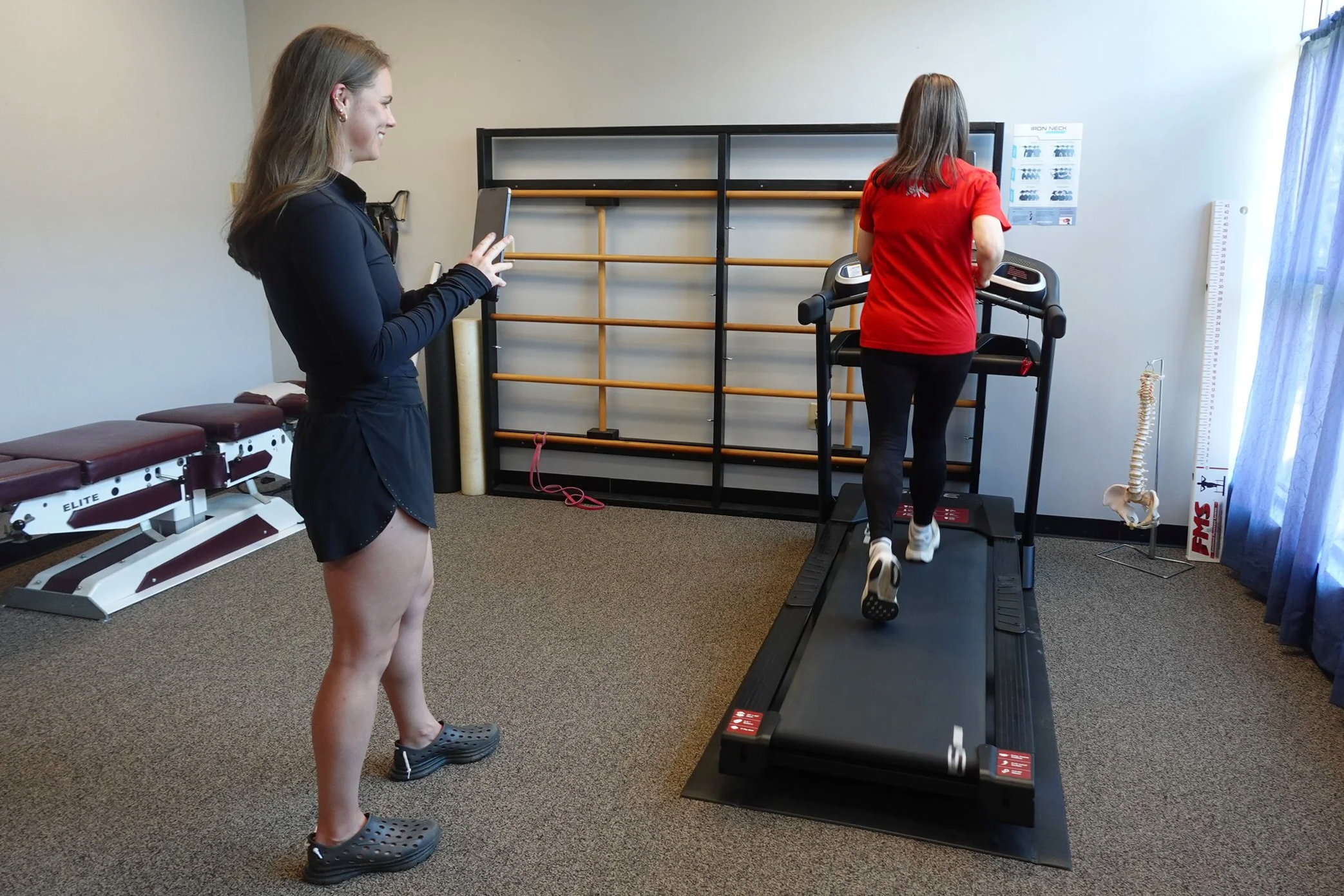 A woman in black athletic clothes taking a photo of another woman wearing a red shirt and black leggings walking on a treadmill. The room has exercise equipment, a spine model, and a height measurement chart.