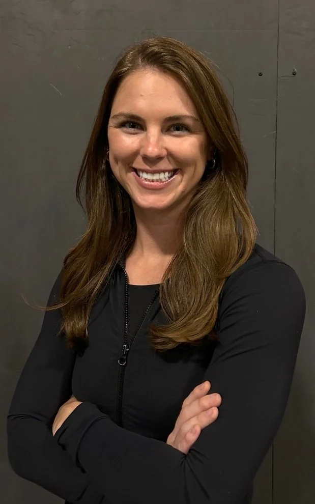 A smiling woman with long brown hair, wearing a black zip-up jacket, standing with her arms crossed against a gray wall background.
