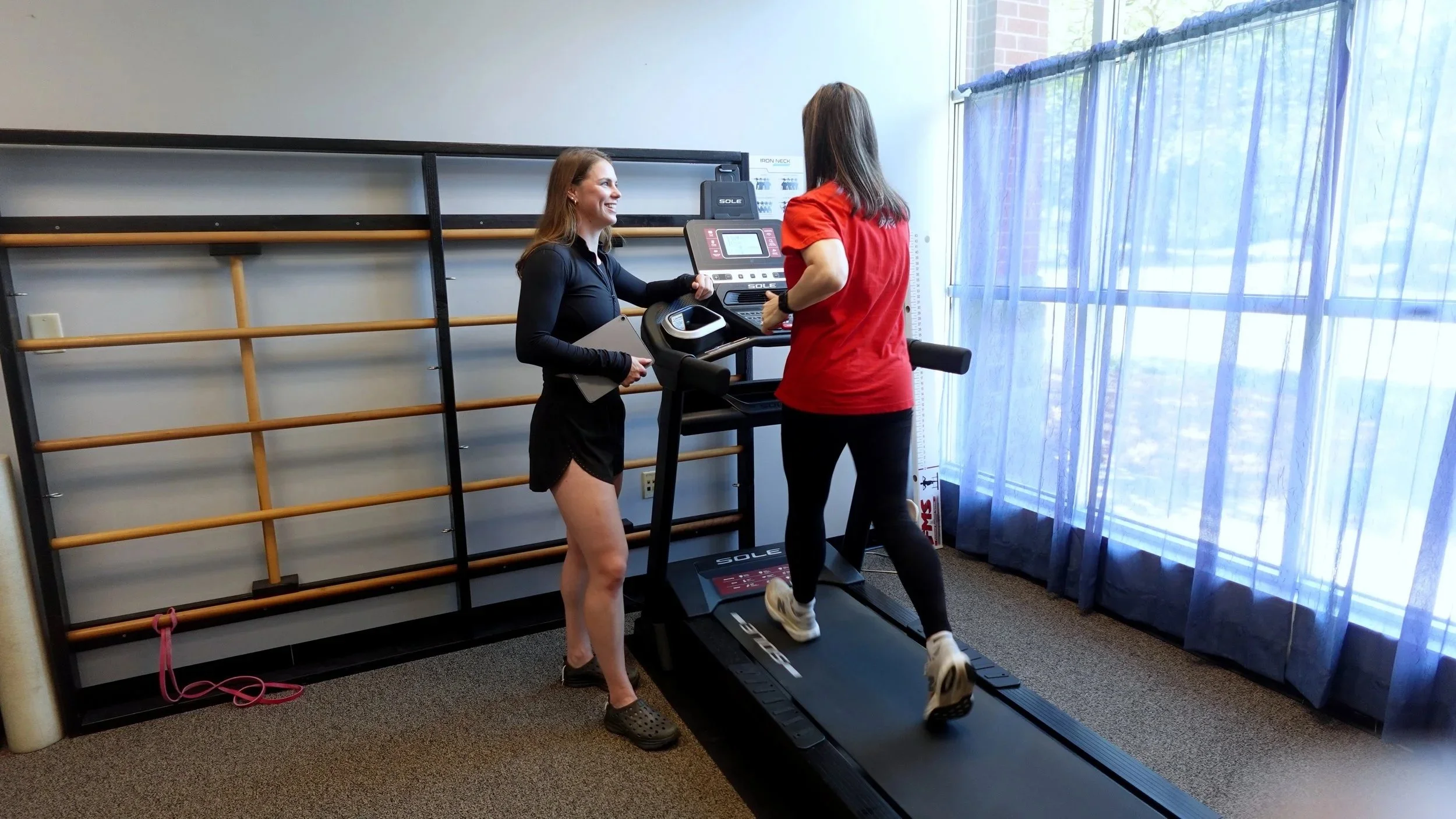 Two women interacting near a treadmill in a room with a large window and blue curtains. One woman is on the treadmill in workout attire, and the other woman, wearing a red shirt, is standing beside her.