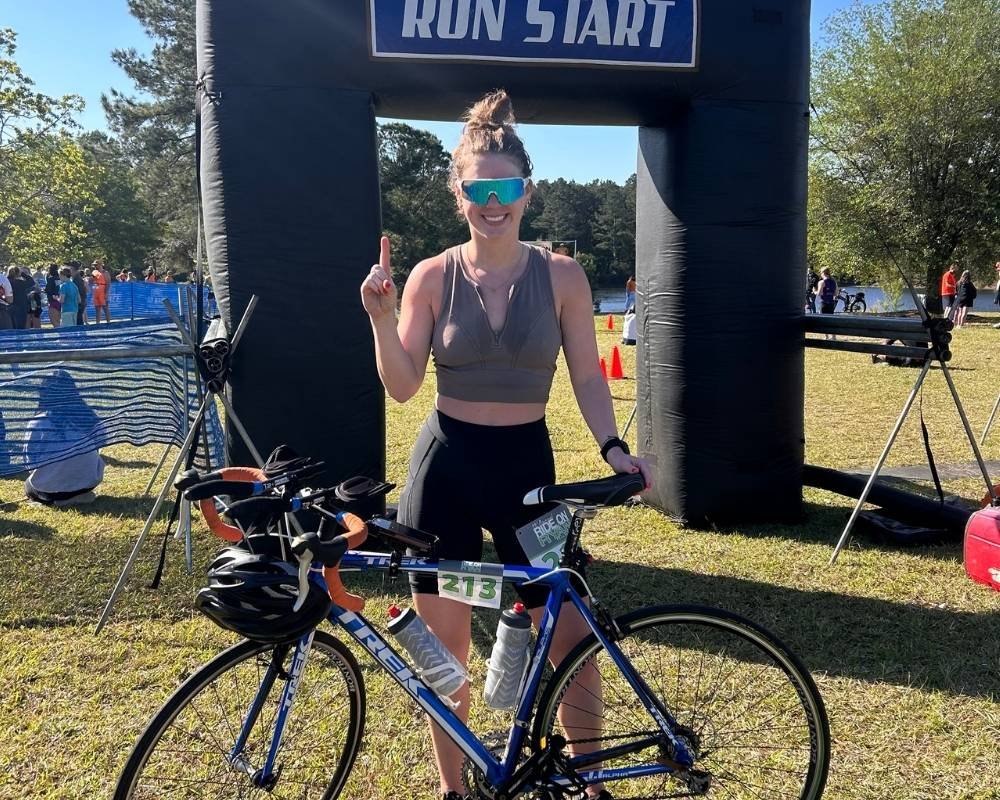 A woman in athletic wear and blue sunglasses is standing at a race start line, holding a blue road bike with a helmet hanging on the handlebar. She is smiling and pointing upward with her right hand. The background shows other participants and spectators, with trees and a river visible in the distance.