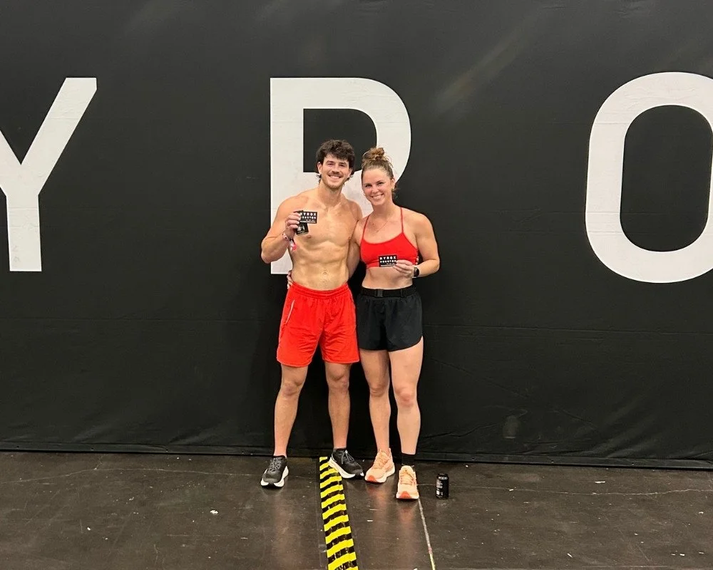 A man and woman in athletic attire standing together in a gym, holding medals, with a black wall behind them that has large white letters. The man is shirtless and wearing red shorts, and the woman is in a red sports bra and black shorts.