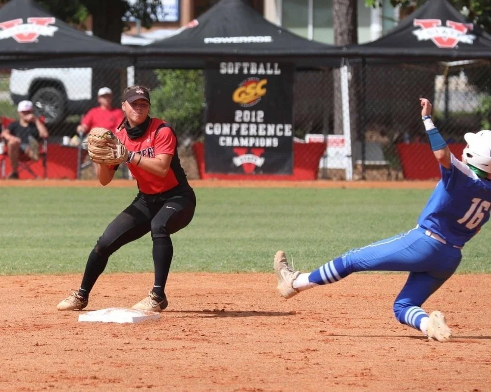 A softball player in a red shirt and black pants is standing on a base, ready to catch a throw with her glove. An opposing player in a blue uniform and white helmet is sliding into the base. The background shows a banner indicating a 2012 conference championship and a chain-link fence with spectators.