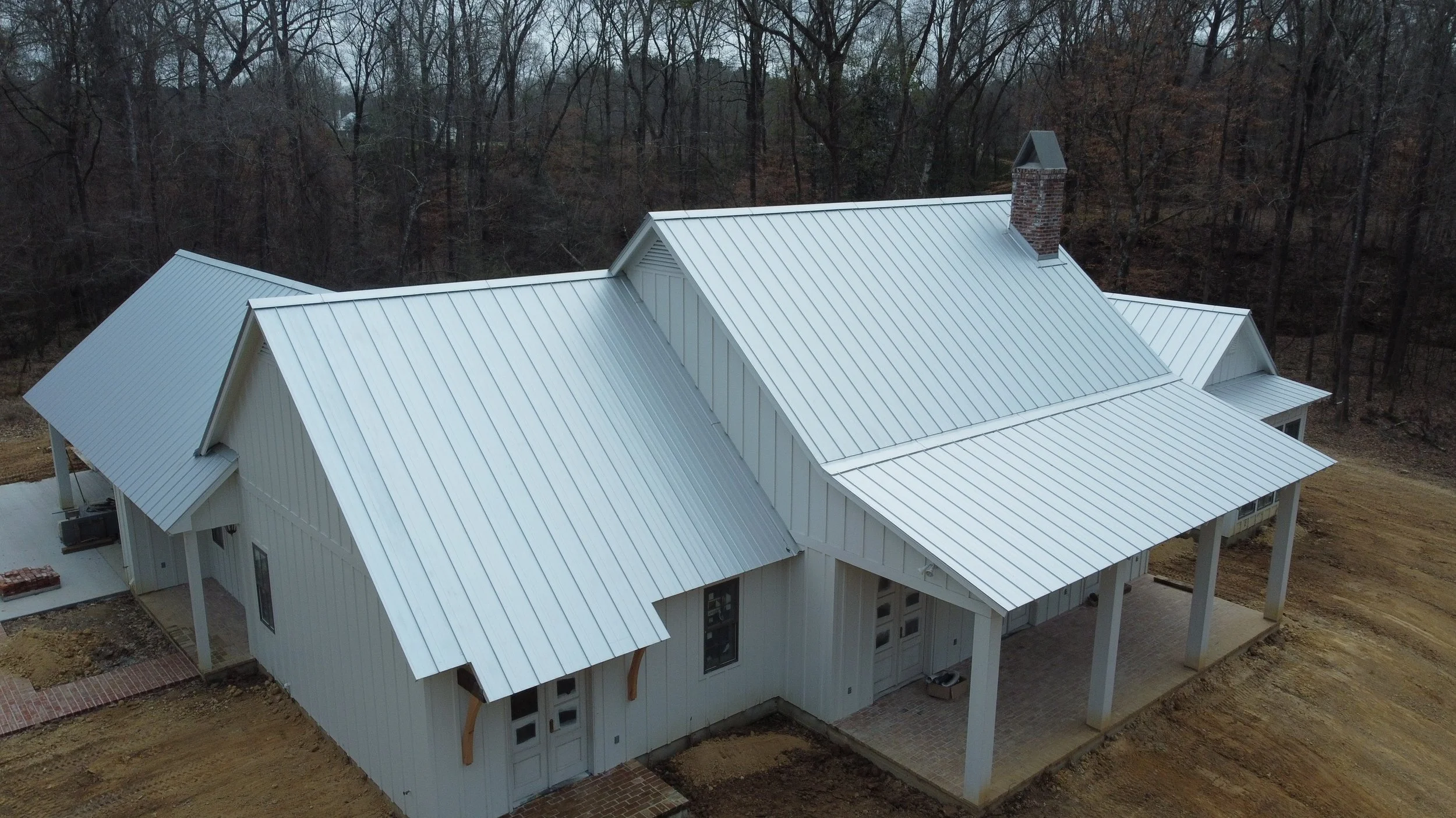 A house with a white metal roof surrounded by trees and a bare lot.