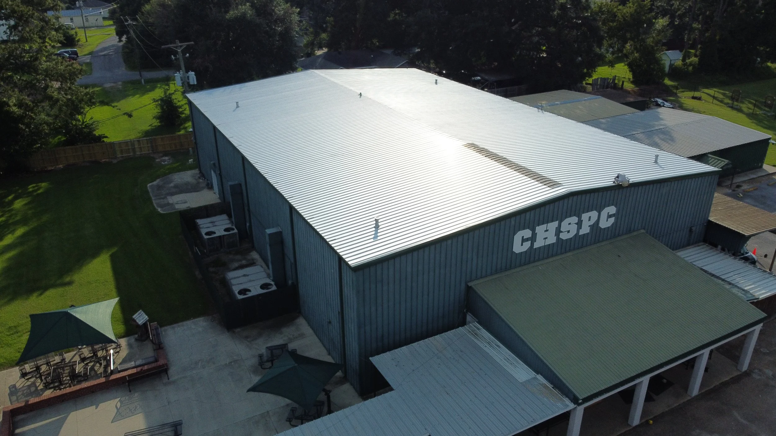 Aerial view of a large industrial building with a metal roof labeled 'CHSPC', surrounded by green lawns, trees, outdoor seating shaded by umbrellas, and nearby residential houses.