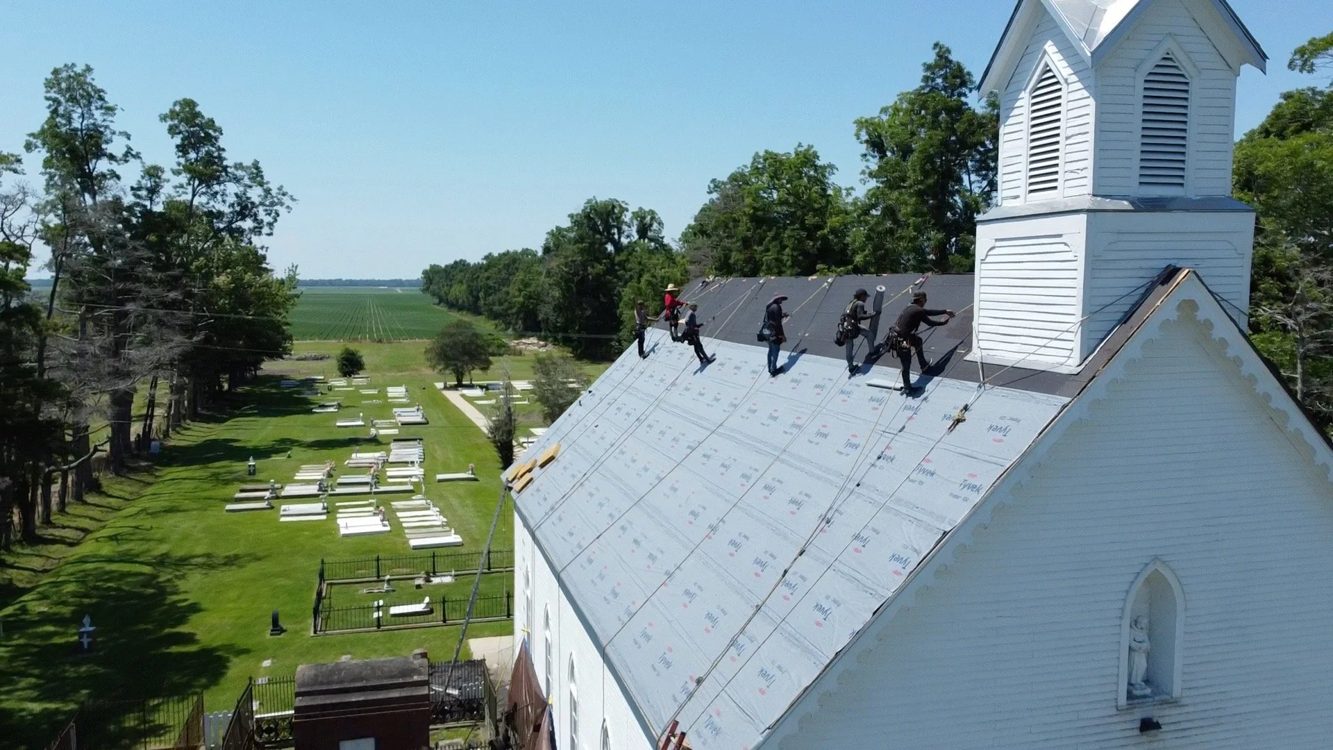 Workers installing or repairing the roof of a white wooden church on a sunny day, with a graveyard and trees in the background.