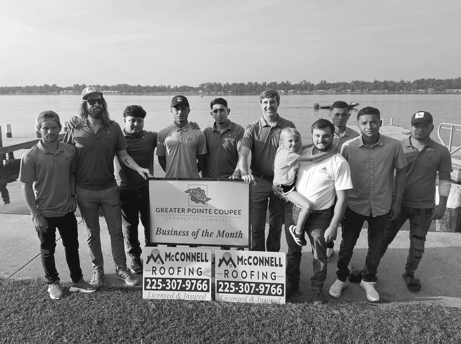 Group of people standing outdoors on a dock beside water, holding a sign that reads 'Greater Pointe Coupee Chamber of Commerce Business of the Month,' with additional signs for McConnell Roofing and contact information.