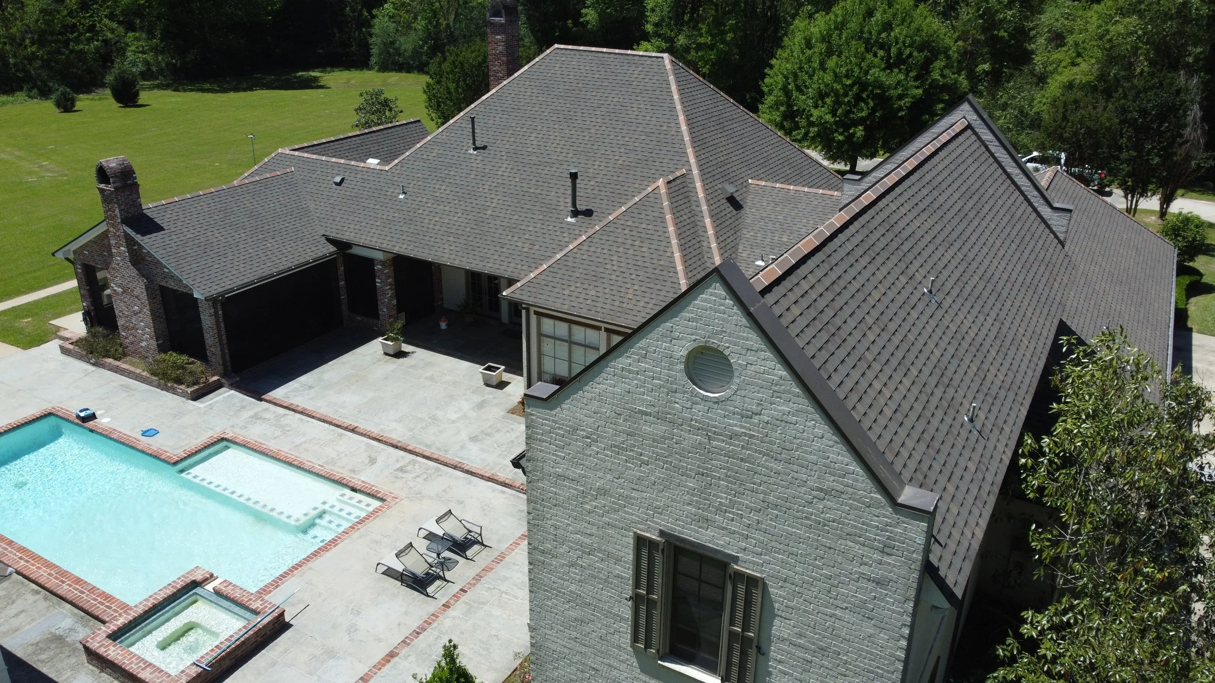 Aerial view of a large house with gray shingle roof, brick chimney, and white brick exterior, adjacent to a swimming pool with lounge chairs and patio area, surrounded by green trees and lawn.