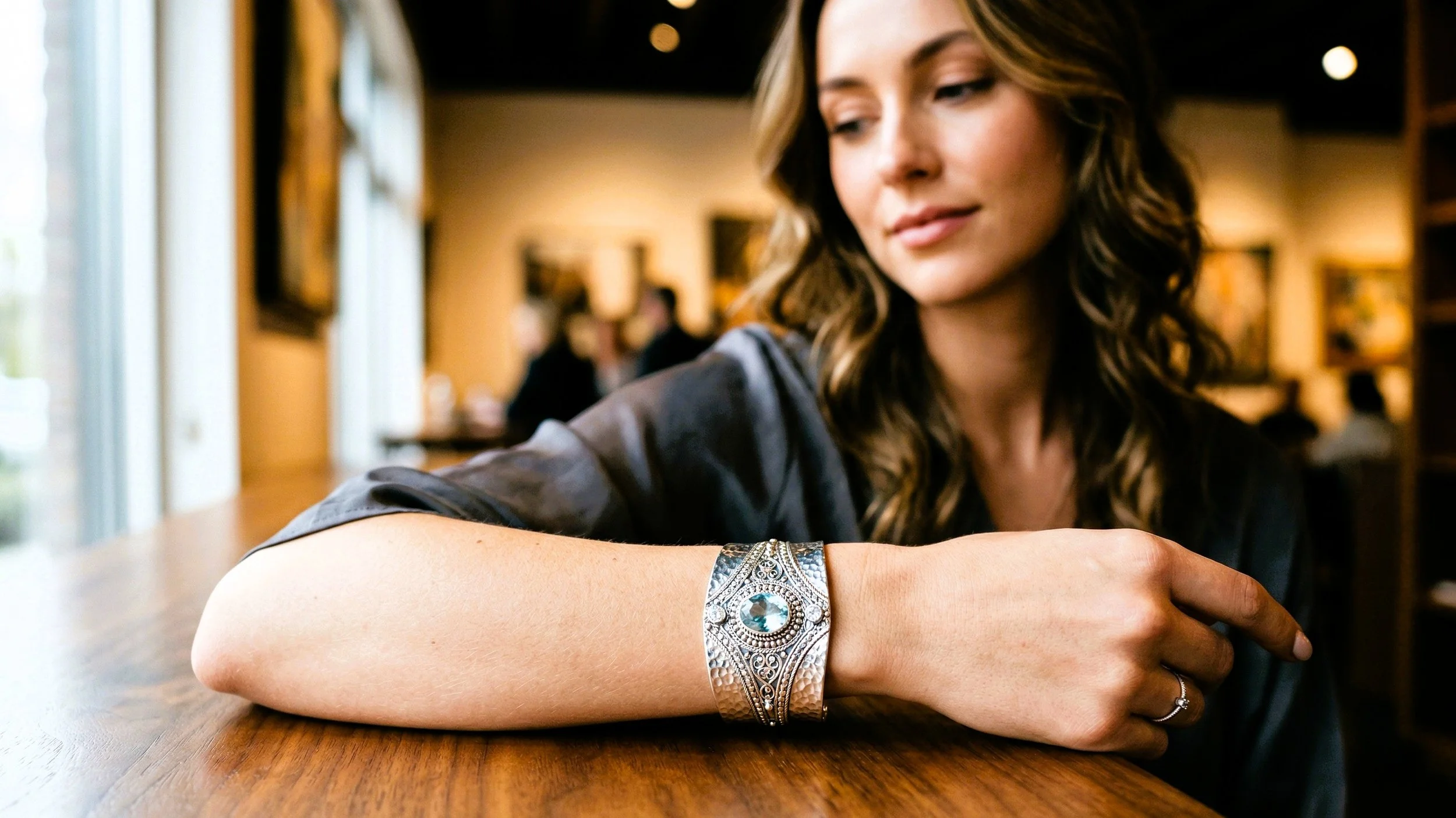 A woman with brunette wavy hair wearing a dark top, sitting at a wooden table in a warmly lit cafe, looking down at her wrist which has a large ornate silver bracelet with a blue gemstone.