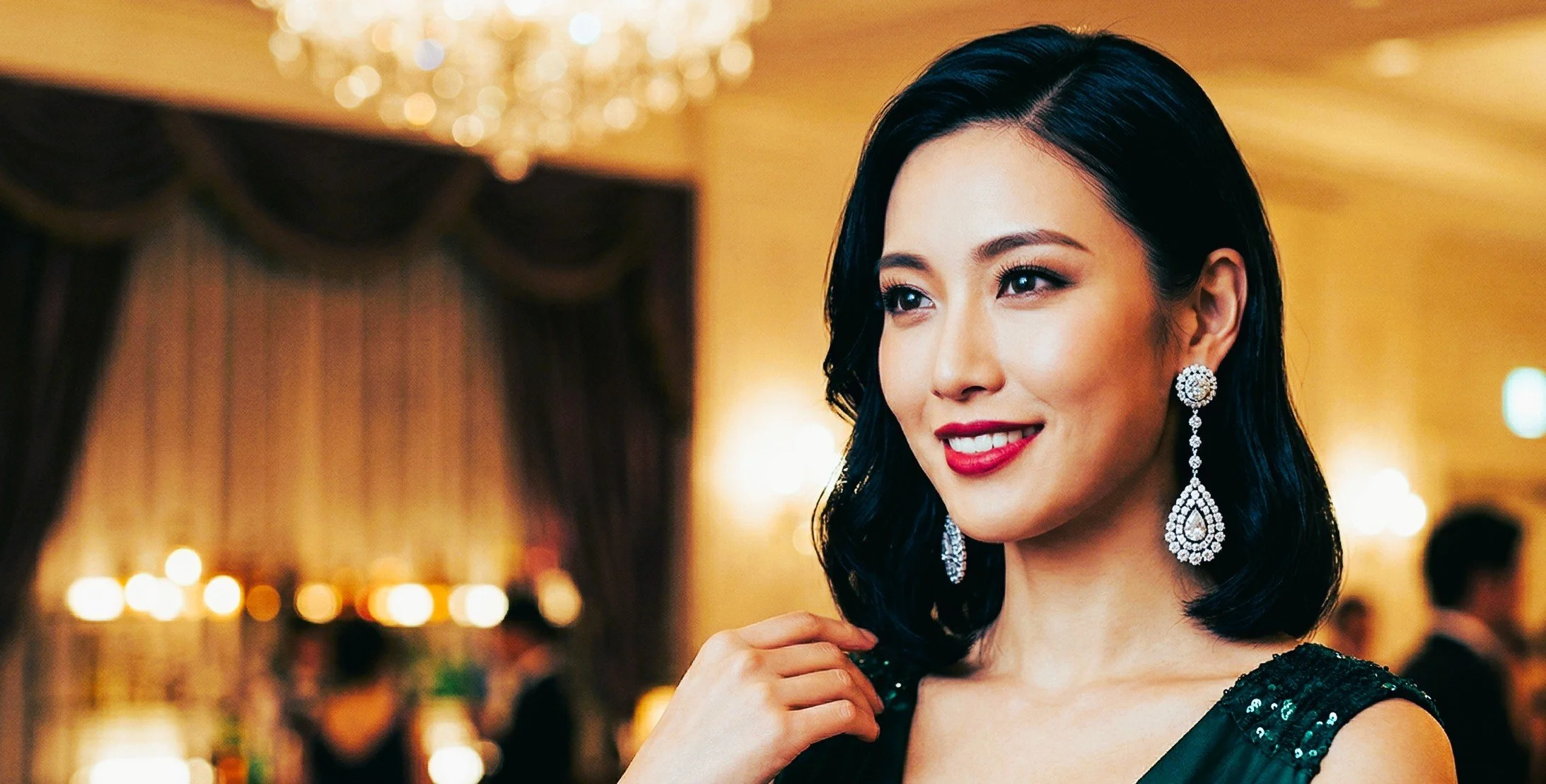 A woman with black hair and makeup, wearing large sparkling earrings and a black sequin dress, smiling at an elegant indoor event.