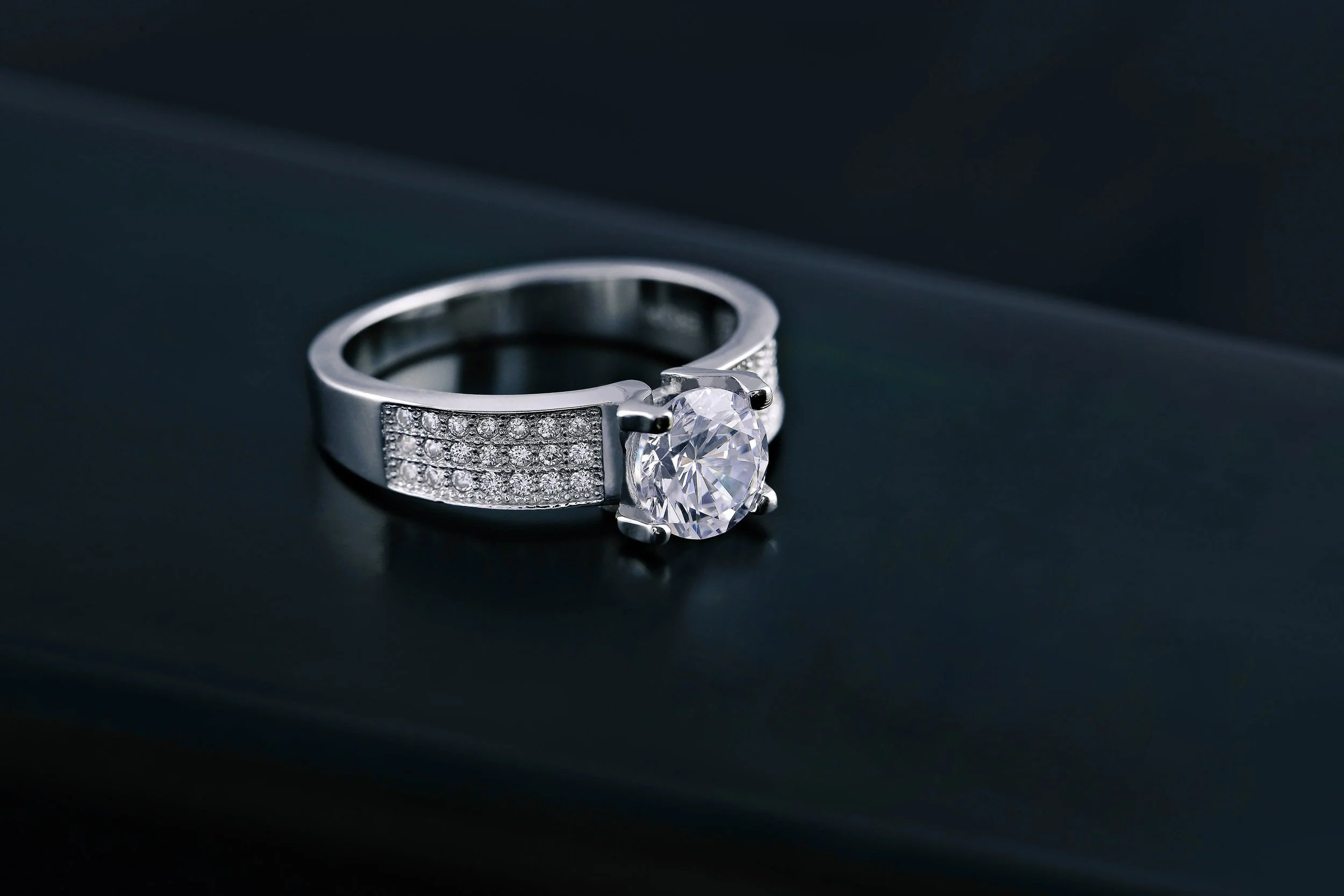 A silver engagement ring with a large round diamond solitaire setting and a band encrusted with small diamonds, placed on a dark surface.