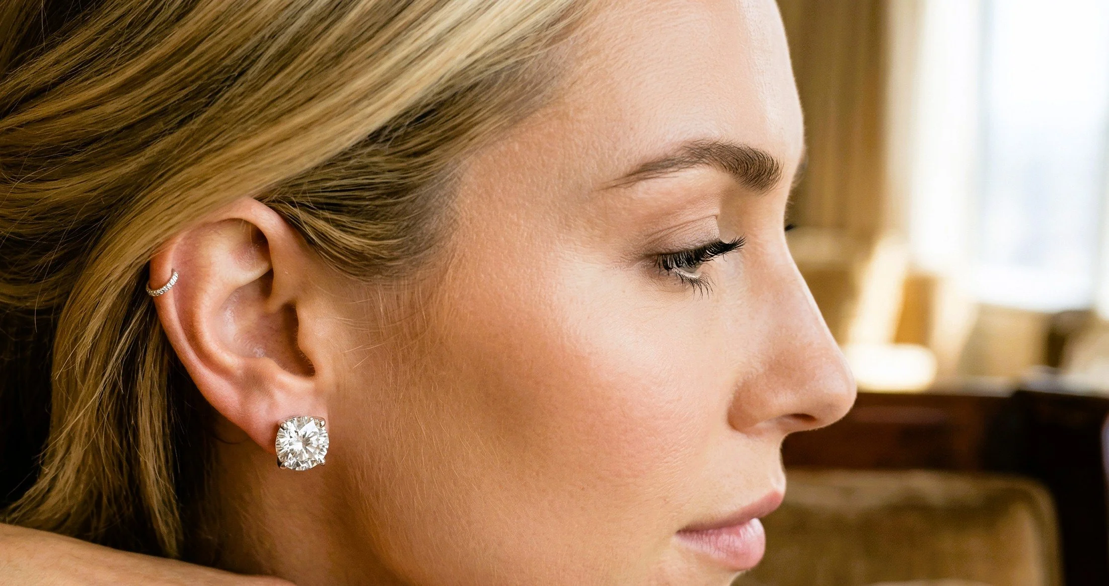 Close-up profile of a woman with blonde hair, wearing diamond jewelry on her ear, with soft natural lighting in a warm indoor setting.