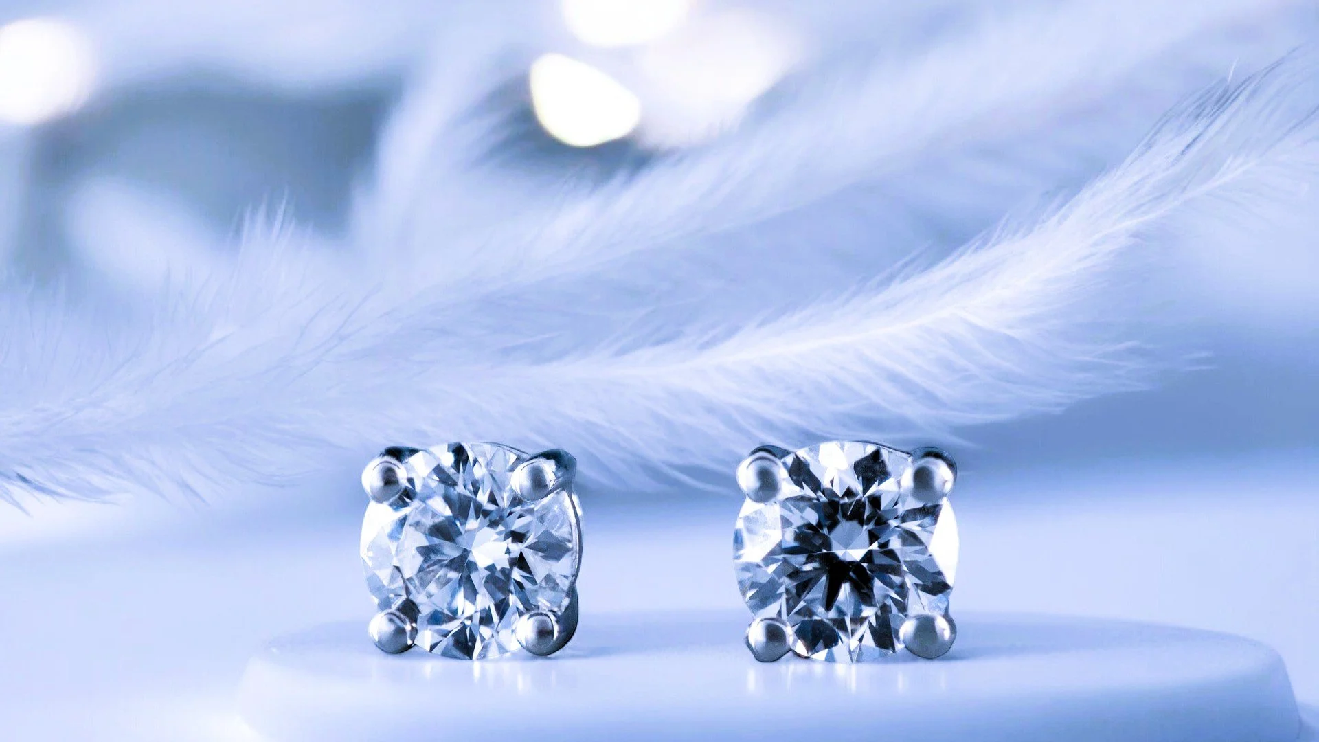 Close-up of a pair of diamond stud earrings resting on a white surface, with light blue feather and soft blurred reflections in the background.